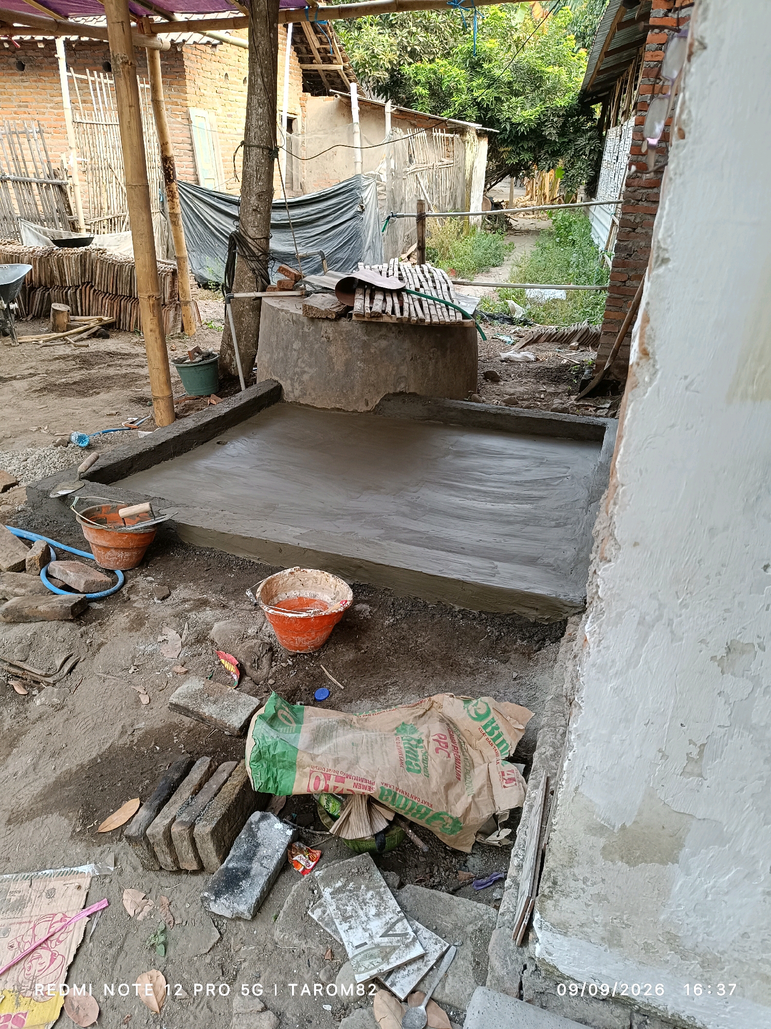 In Rejoagung, Indonesia, an outdoor scene depicts the construction of a concrete floor. The foreground is dominated by a wet, gray concrete surface, meticulously leveled and smoothed, bordered by a newly formed concrete edge. Two orange buckets, one containing water and the other appearing empty, sit nearby, along with scattered bricks and a blue hose. In the mid-ground, a large, rough concrete structure, possibly a well or cistern, is visible. Beyond this, a collection of stacked ceramic tiles suggests ongoing or recent work. The background reveals a rustic environment with wooden poles supporting a thatched roof, a brick building, and overgrown vegetation, hinting at a rural or semi-rural setting. The lighting suggests it is late afternoon, with a timestamp at the bottom right corner indicating the date and time as 09/09/2026, 16:37. A watermark at the bottom left reads "REDMI NOTE 12 PRO 5G | TAROM82".