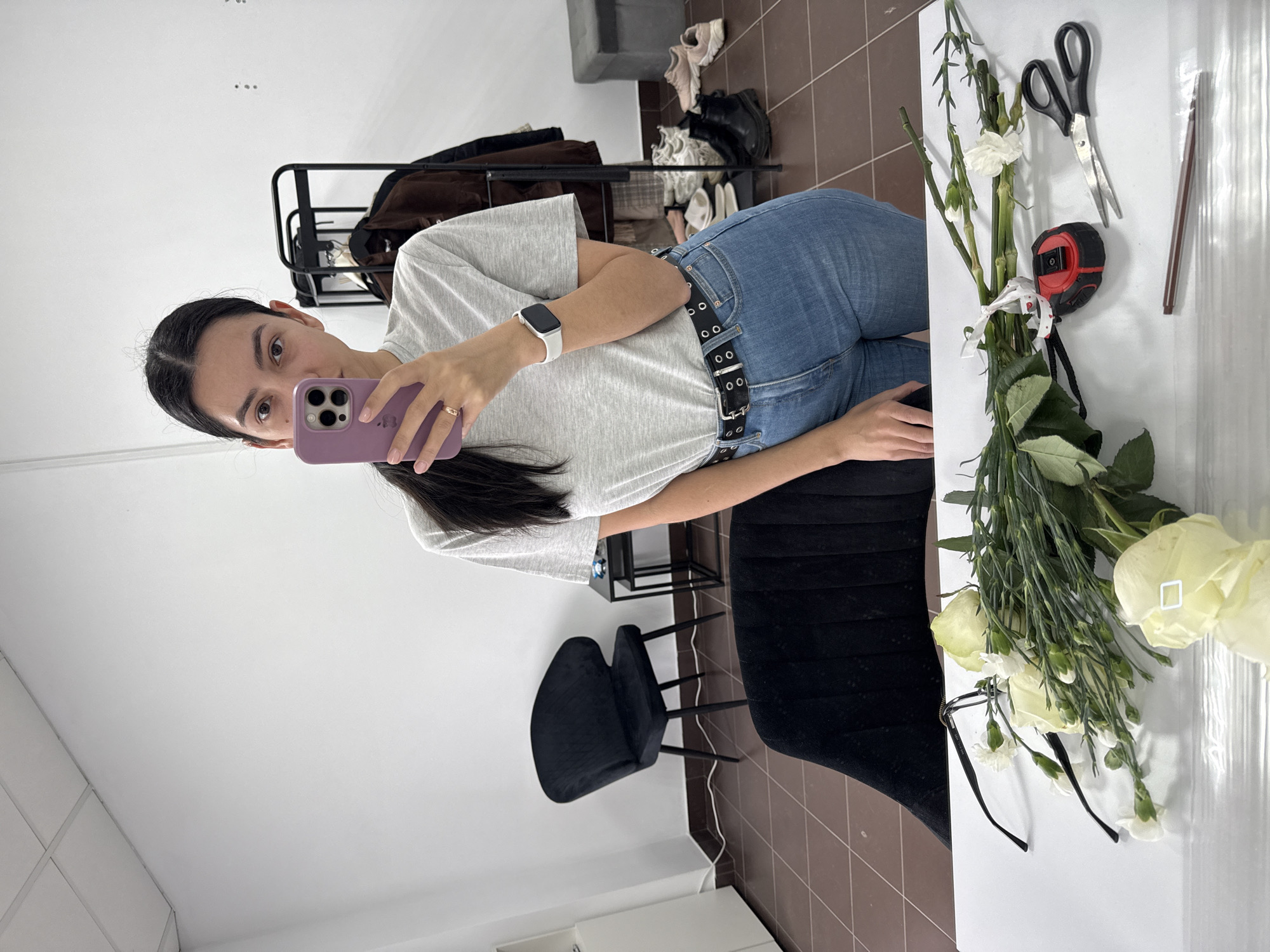 A young woman is captured in a mirror selfie, looking directly at her purple smartphone, which features three camera lenses and is held in her right hand. She has dark hair and wears a light grey t-shirt, blue jeans, and a black belt adorned with silver eyelets. A white smart watch is on her left wrist, and a gold ring is on her right ring finger.
In front of her, on a white surface, lies a bouquet of white flowers, including roses and what appear to be white carnations, along with green foliage. Nearby are black-handled scissors, a red and black measuring tape, and a pair of black-framed glasses. A transparent ruler-like object is also visible on the surface.
The background reveals an indoor setting with white walls and a brown tiled floor. To the woman's left, a black metal clothing rack holds various garments, including a brown jacket. Several pairs of shoes, such as sneakers, boots, and sandals, are scattered on the floor beneath the rack. To her right, a black upholstered chair is partially visible. The lighting is bright and uniform, suggesting a daytime setting, possibly illuminated by artificial light. The overall scene implies an activity involving flowers, potentially in a studio, dressing room, or retail space. No text is visible in the image.