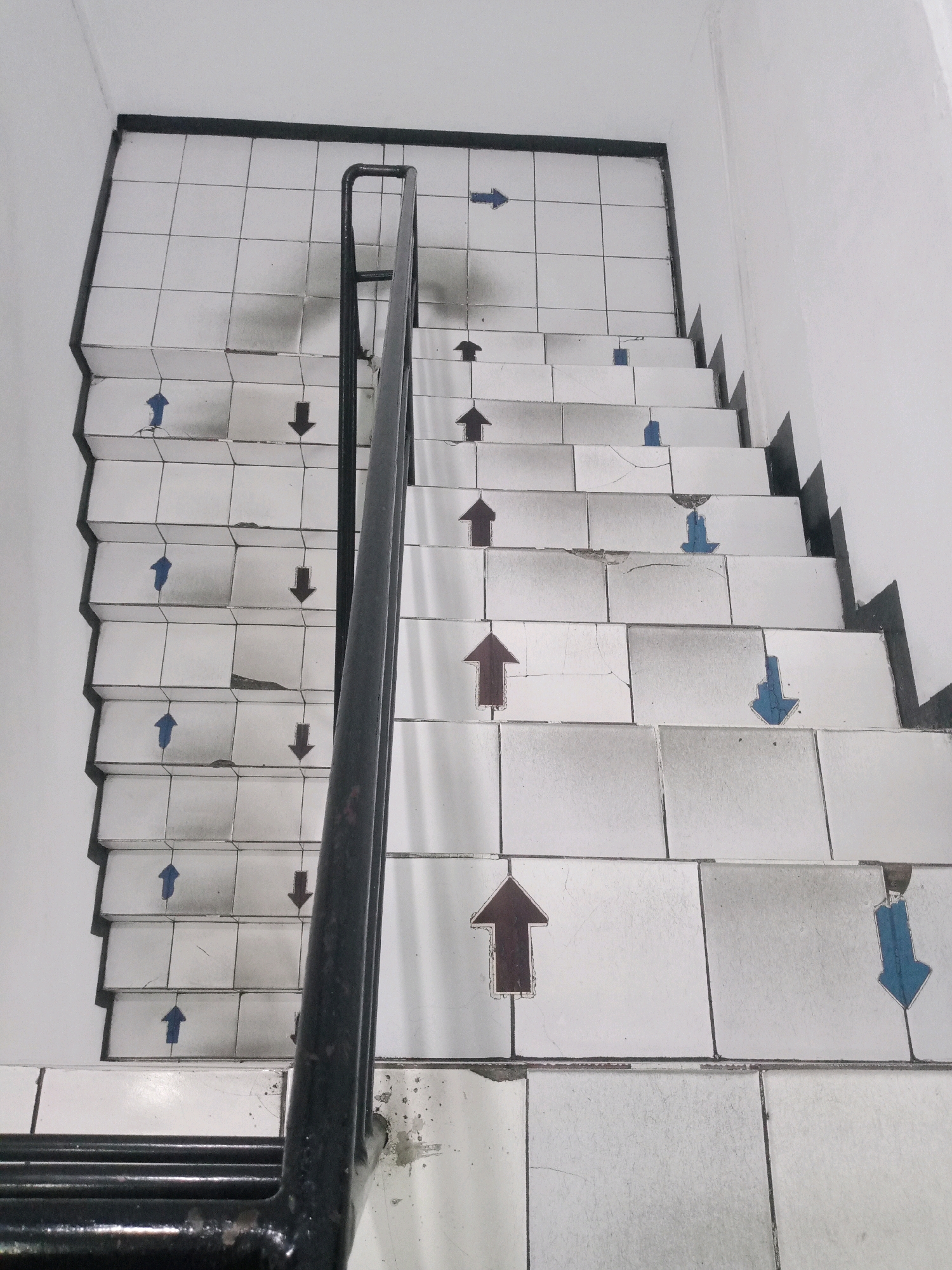 The image shows a tiled staircase in an indoor environment. The tiles are white with dark grout lines. Each step has an arrow painted on it, indicating direction. Some arrows are brown and point upwards, while others are blue and point downwards. The handrail is black and appears to be made of metal. The walls surrounding the staircase are white. The overall scene suggests a public or institutional building. The location is described as being in Balaraja, Indonesia.