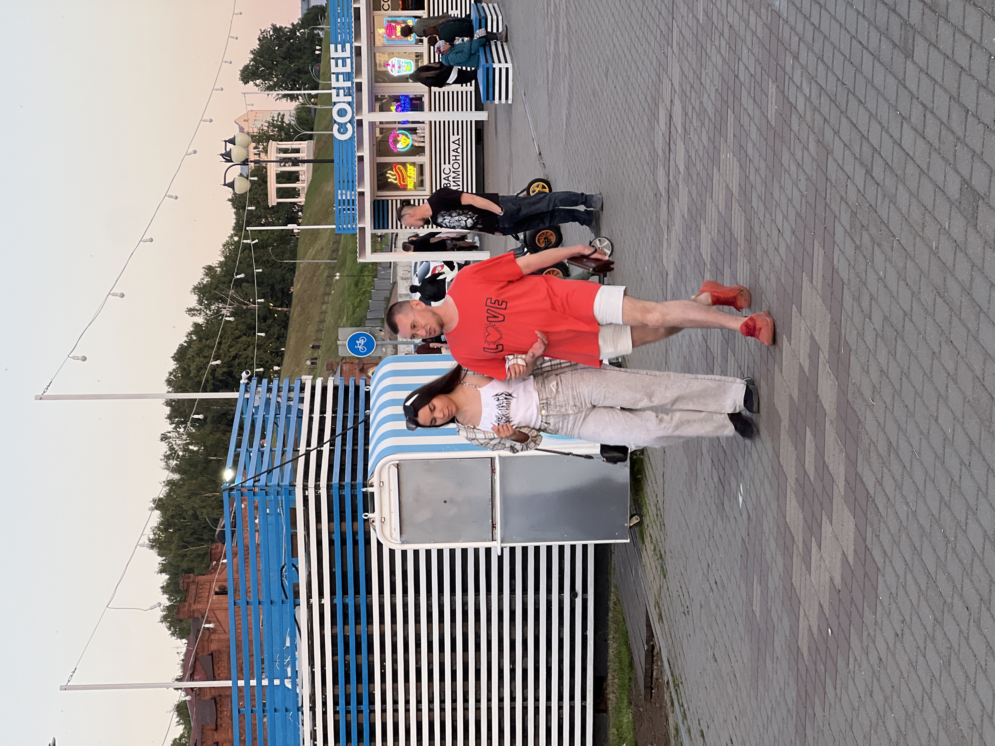 This image depicts a late afternoon or early evening scene on a paved urban promenade, specifically identified as the Izhevsk Pond embankment in Izhevsk, Russia. The sky is a gradient of pale blue to light pink, suggesting dusk.
In the foreground, a woman and a man walk from left to right. The woman has long dark hair, wears sunglasses on her head, a white tank top with a dark graphic, light-colored wide-leg pants, and black shoes, holding a white cup in her left hand. The man, walking to her right, has short brown hair, wears glasses, a red t-shirt with "LOVE" printed in white, white shorts, and red sneakers. He holds a dark object, possibly a phone or wallet, in his left hand.
The promenade is paved with gray and reddish-brown interlocking pavers. To the left, a large, multi-tiered structure built from horizontal blue and white slats stands, with a smaller, gray mobile kiosk positioned beside it. To the right, a prominent "COFFEE" stall features a white frame, blue accents, and multiple neon signs depicting items like a hot dog, drinks, and "ВАС ЛИМОНАД" (YOUR LEMONADE) in Cyrillic. Several people are visible near this stall; one man in a black t-shirt and jeans stands beside a yellow child's pedal car, while others are seated on blue and white striped benches.
Overhead, string lights with exposed bulbs are strung across the scene. A blue circular sign with a white bicycle icon is visible to the left of the coffee stall, indicating a bicycle path. In the background, a lush green hillside is covered with trees, topped by a white gazebo. Further to the left, older red brick buildings with dark roofs are partially visible behind the hillside vegetation.