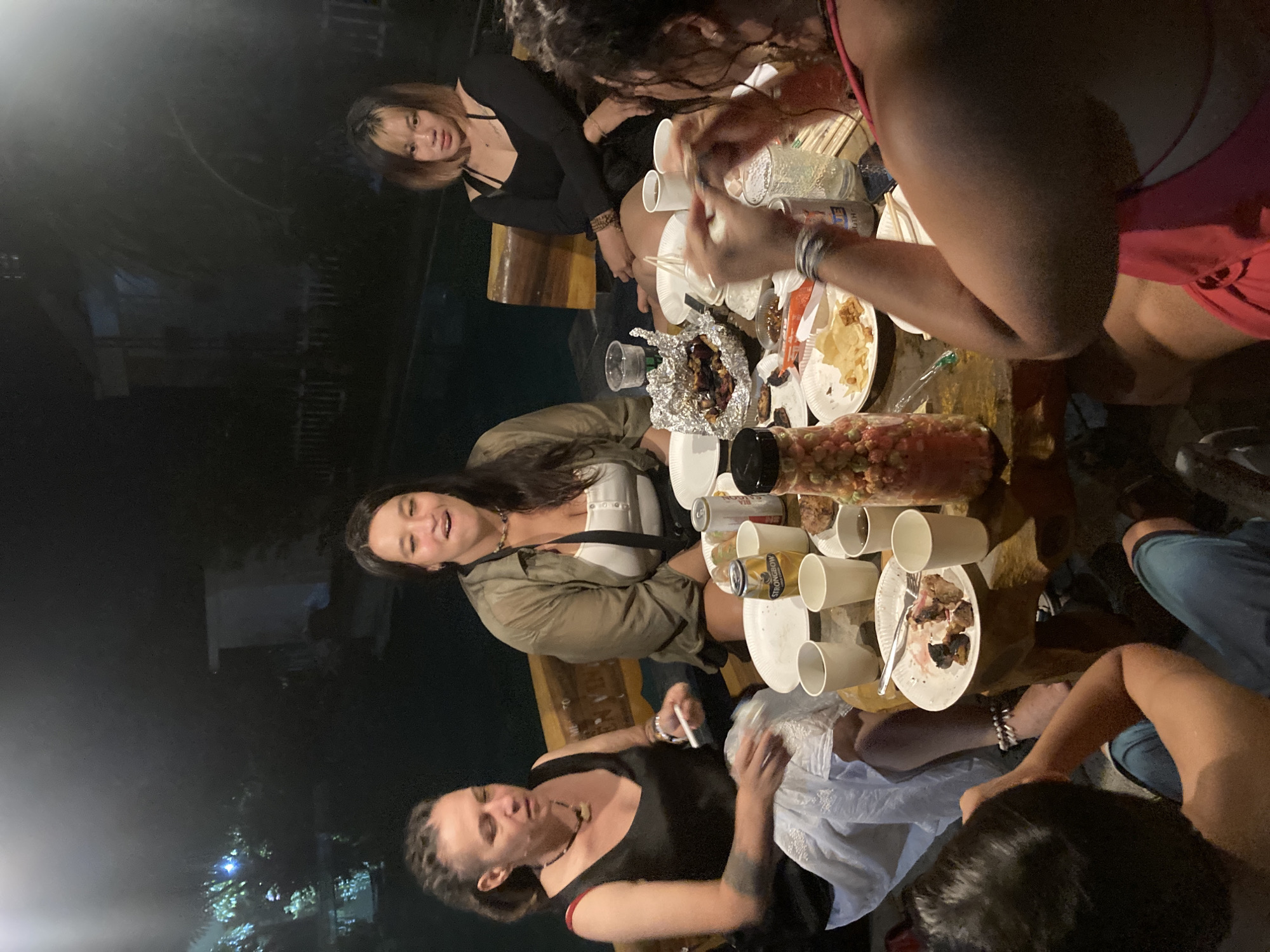 A nighttime gathering takes place in Mũi Né, Vietnam. Six women are around a wooden table laden with food, cups, and cans. One woman is holding a cigarette, and the table contains plates of partially consumed food.