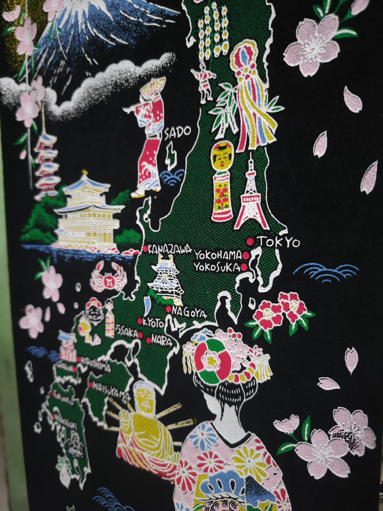 The image is a close-up, vertical, medium shot of a black fabric tapestry depicting a stylized map of Japan. The map is rendered in green with white outlines, and it is adorned with various illustrations of Japanese landmarks, cultural elements, and natural scenery. Notable depictions include Mount Fuji in the upper left, a pagoda, a person in traditional Japanese attire, cherry blossoms, a crab, and a representation of the Eiffel Tower. Several Japanese cities are labeled in white text, such as SADO, KANAZAWA, YOKOHAMA, YOKOSUKA, TOKYO, NAGOYA, KYOTO, OSAKA, NARA, HIROSHIMA, MATSUYAMA, and NAGASAKI. The tapestry also features illustrations of a person in a kimono with an elaborate floral headdress, and a seated Buddha figure. The overall style is colorful and illustrative, with a focus on iconic Japanese imagery. There is no visible presence of Sroyo, Indonesia, in this image.