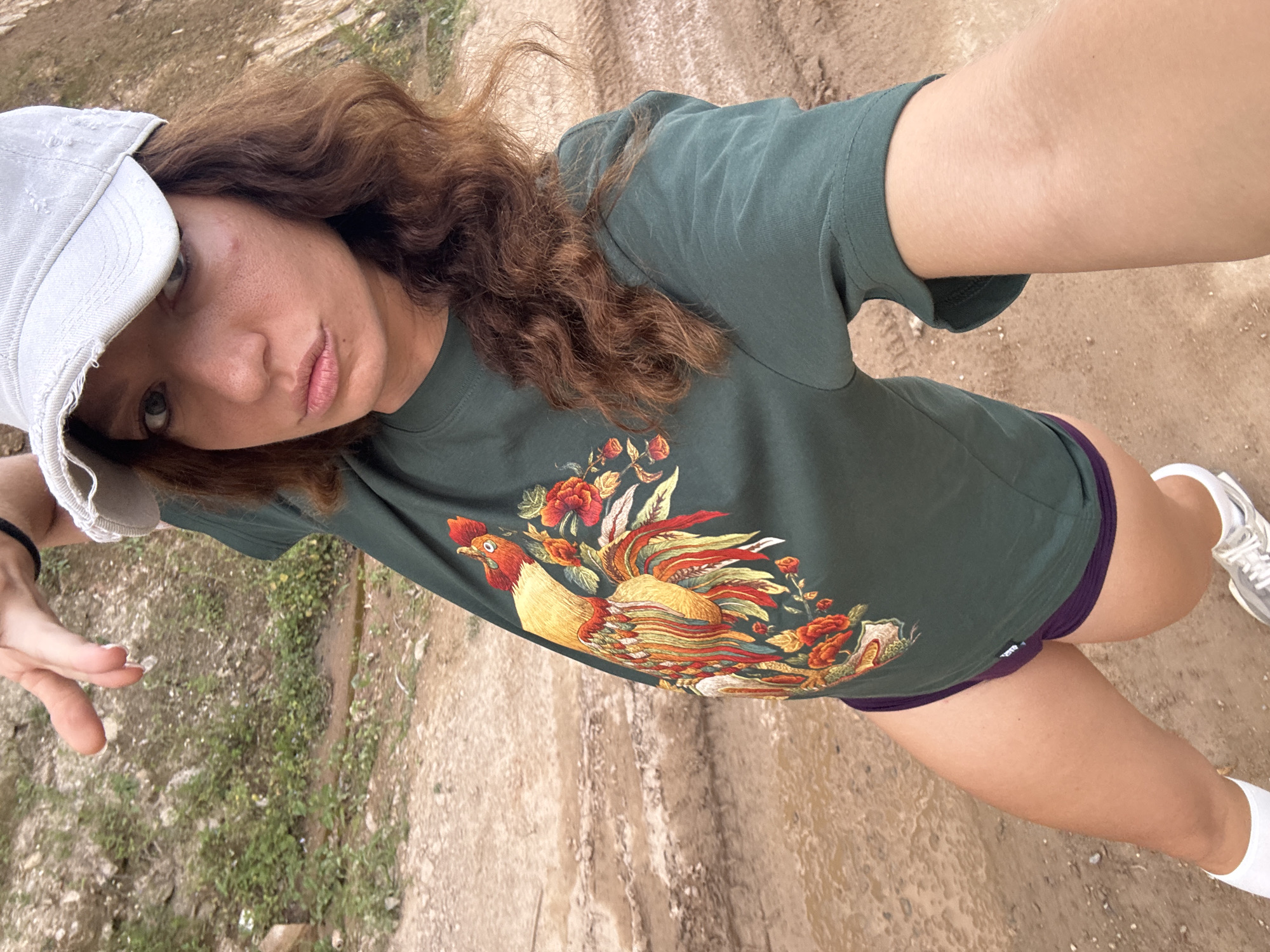 A young woman is standing on a dirt road, possibly in a rural area within Pờ Ly Ngài Commune, Vietnam. She is taking a selfie, holding the camera in her outstretched hand. She wears a green t-shirt with a rooster design, purple shorts, white socks, and white sneakers. She also has a light-colored baseball cap on her head and her curly hair is loose. The dirt road has tracks and appears to be muddy in places. There's some vegetation visible to the left of the road.
