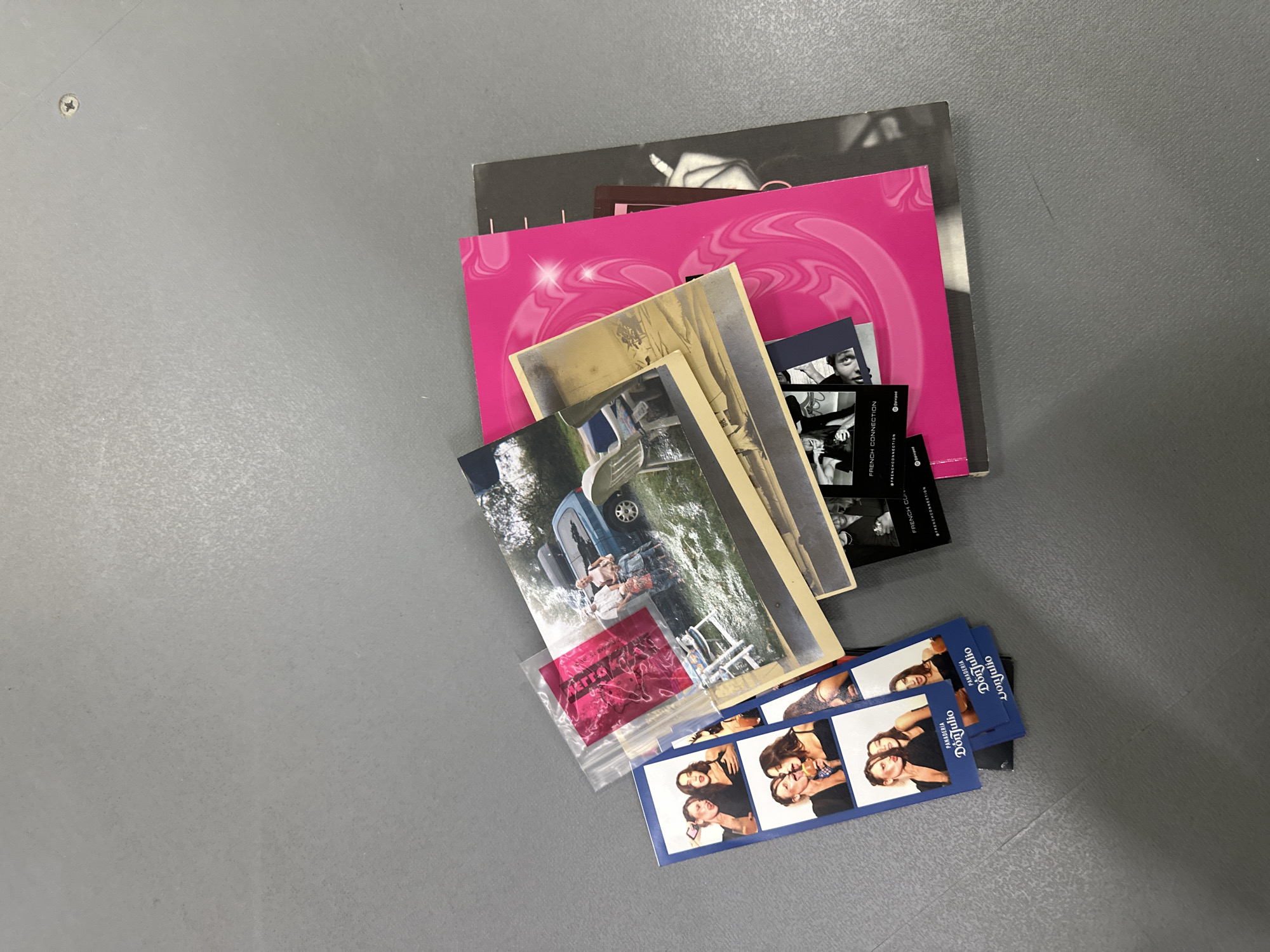 The image shows a collection of items scattered on a gray surface. The items include a pink book or folder, a black and white book or folder, several photographs, including one depicting a blue car, and strips of photobooth-style pictures of two women. One photograph is in a plastic bag. The words "DON JULIO" are visible on the photo booth strips. The location is not discernible from the image.