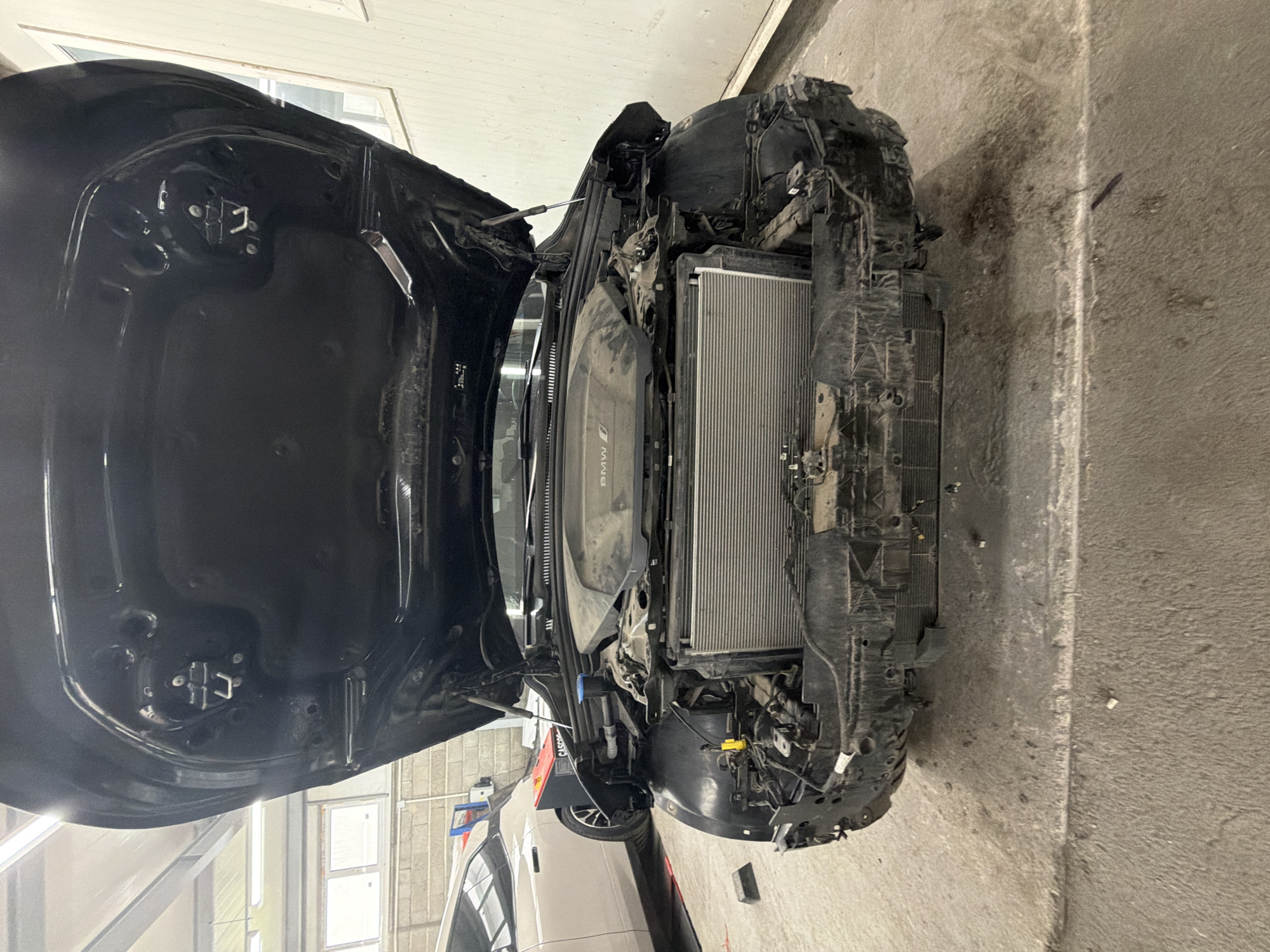 An open black car hood reveals the engine bay and a large radiator at the front of the vehicle. The car appears to be in a workshop or garage. The floor is concrete and dusty. In the background, another car is partially visible, and there are windows and walls that suggest an indoor industrial setting. The image captures the front section of the car with its bumper removed, exposing internal components.