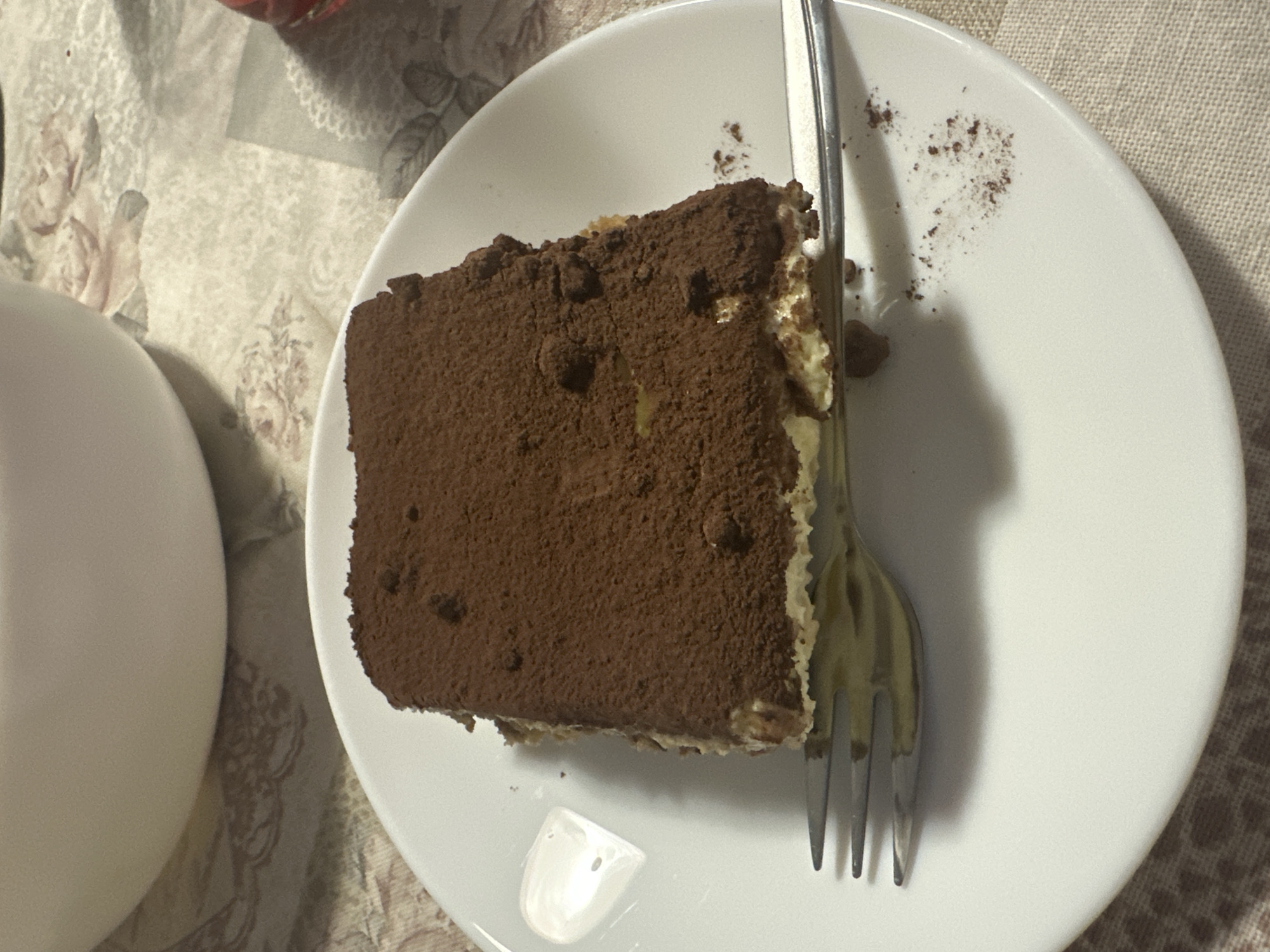 A slice of tiramisu rests on a white plate alongside a fork. Some cocoa powder is sprinkled on the plate. A white ceramic object is visible to the left of the plate. The plate is on a surface with a patterned cloth. The image was likely taken indoors. There is no indication that this image was taken in Varna, Bulgaria.