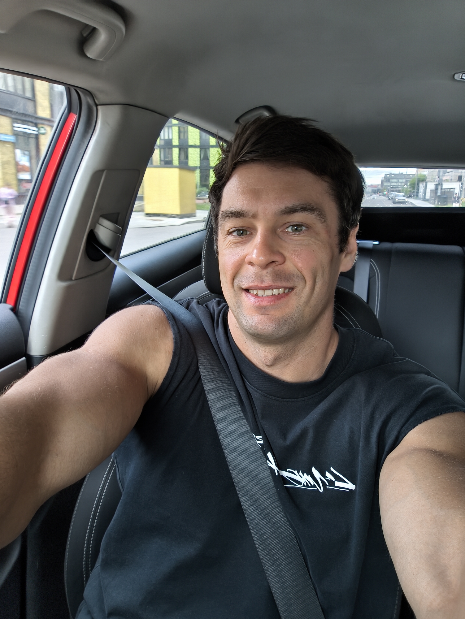 A man is taking a selfie in the driver's seat of a car. He is wearing a black t-shirt and a seatbelt. Through the car windows, a city street with buildings and other cars is visible. It is not possible to confirm the location as Moscow, Russia from the image.