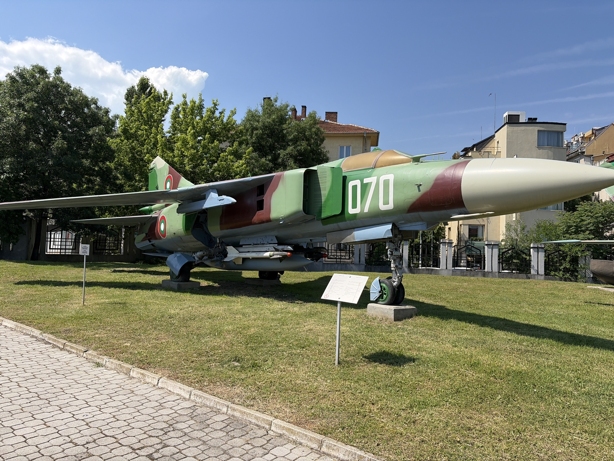 This is a photograph of a camouflaged military jet aircraft, specifically a MiG-21, on display outdoors in the city of Sofia, Bulgaria. The aircraft is painted in green and brown camouflage with white numbers "070" on its fuselage. It is positioned on a grassy area with a paved walkway in the foreground. In the background, there are buildings, trees, and a clear blue sky with some clouds. The jet is on display, likely in a museum or memorial park, indicated by the presence of information plaques near the aircraft. No people are visible in the image.