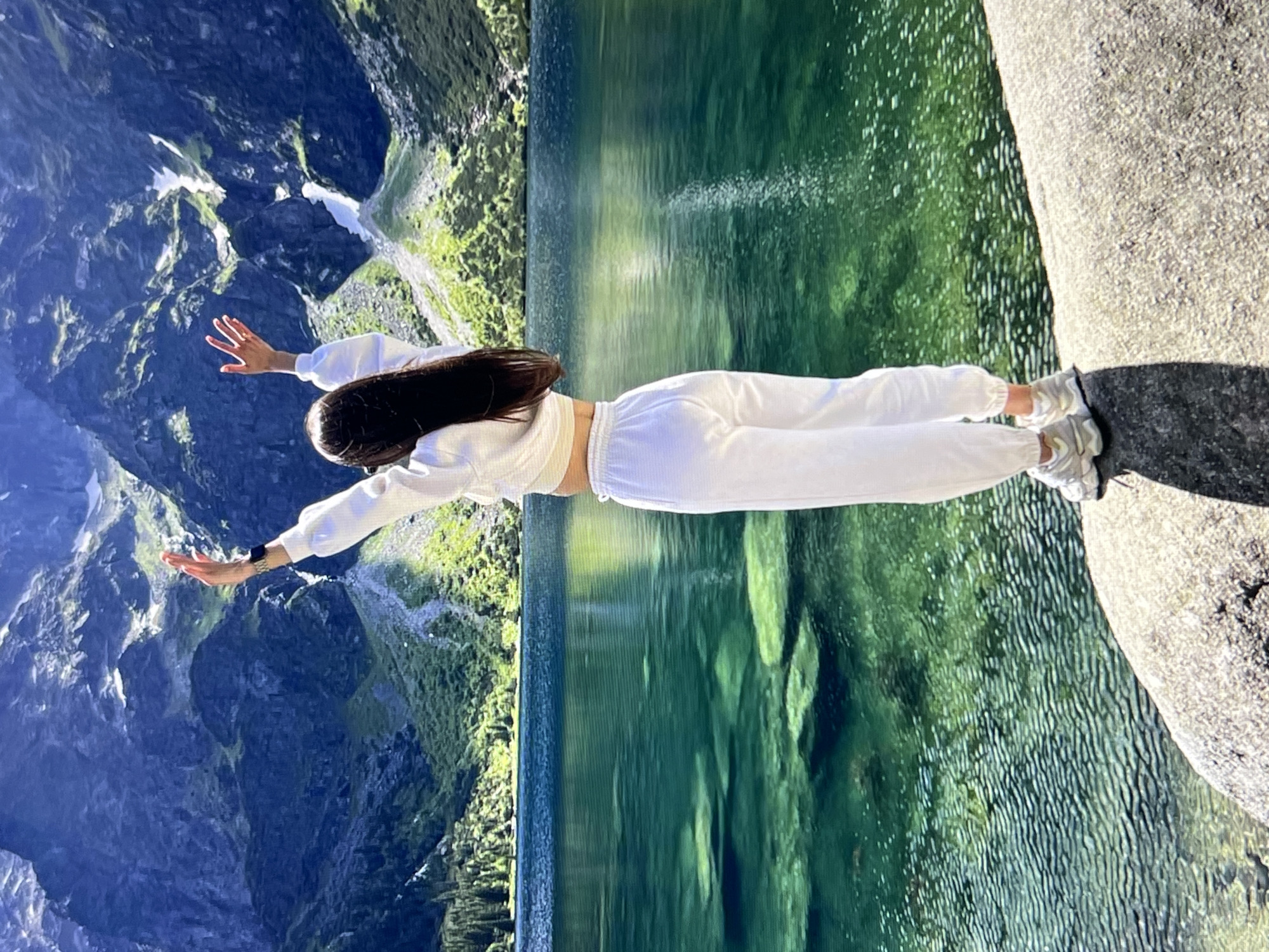 A young woman with long, dark hair stands on a rocky shore, facing away from the camera with her arms raised in a gesture of exhilaration. She is wearing a white cropped hoodie and white sweatpants. The background is a breathtaking natural landscape featuring a clear, emerald-green lake with visible rocks beneath the surface, and towering, rugged mountains that are partially covered in snow and lush green vegetation. The sun is shining, casting a warm light on the scene.