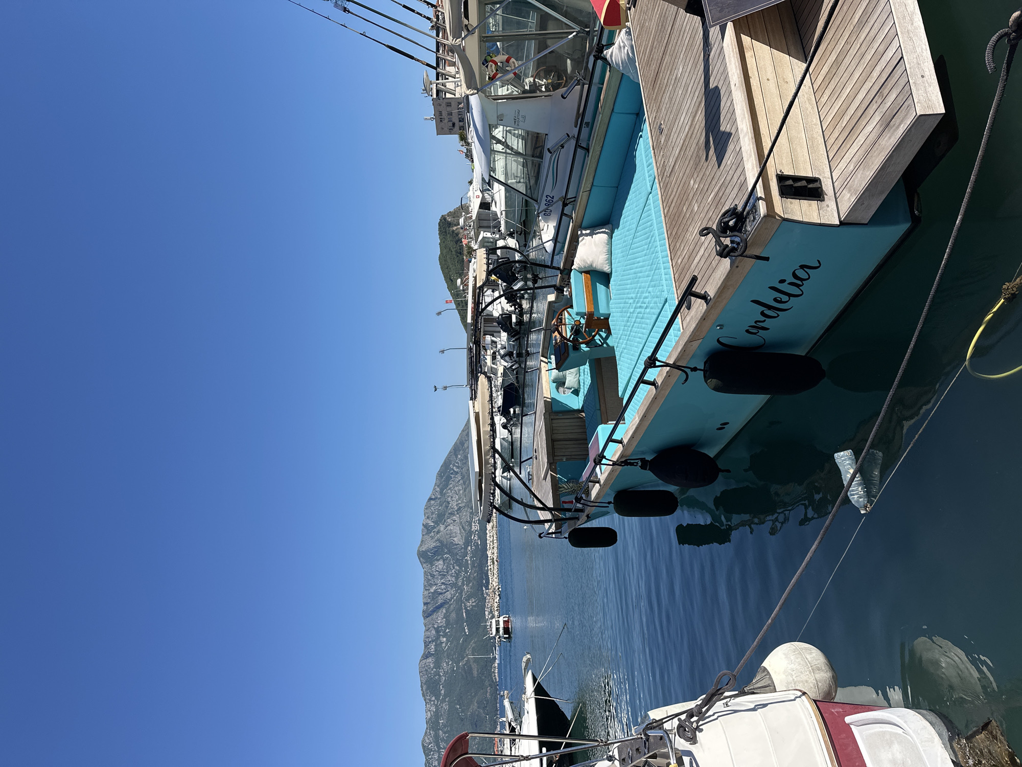 A bright, sunlit scene captured in Budva, Montenegro, features the stern of a docked yacht named "Cordelia." The yacht has a turquoise hull and a wooden deck. Black fenders hang along the side. The clear, blue water reflects the yacht and surroundings. Mountains rise in the background, partially obscuring the city of Budva. Other boats are docked nearby, their structures and rigging partially visible. The sky is clear and blue.