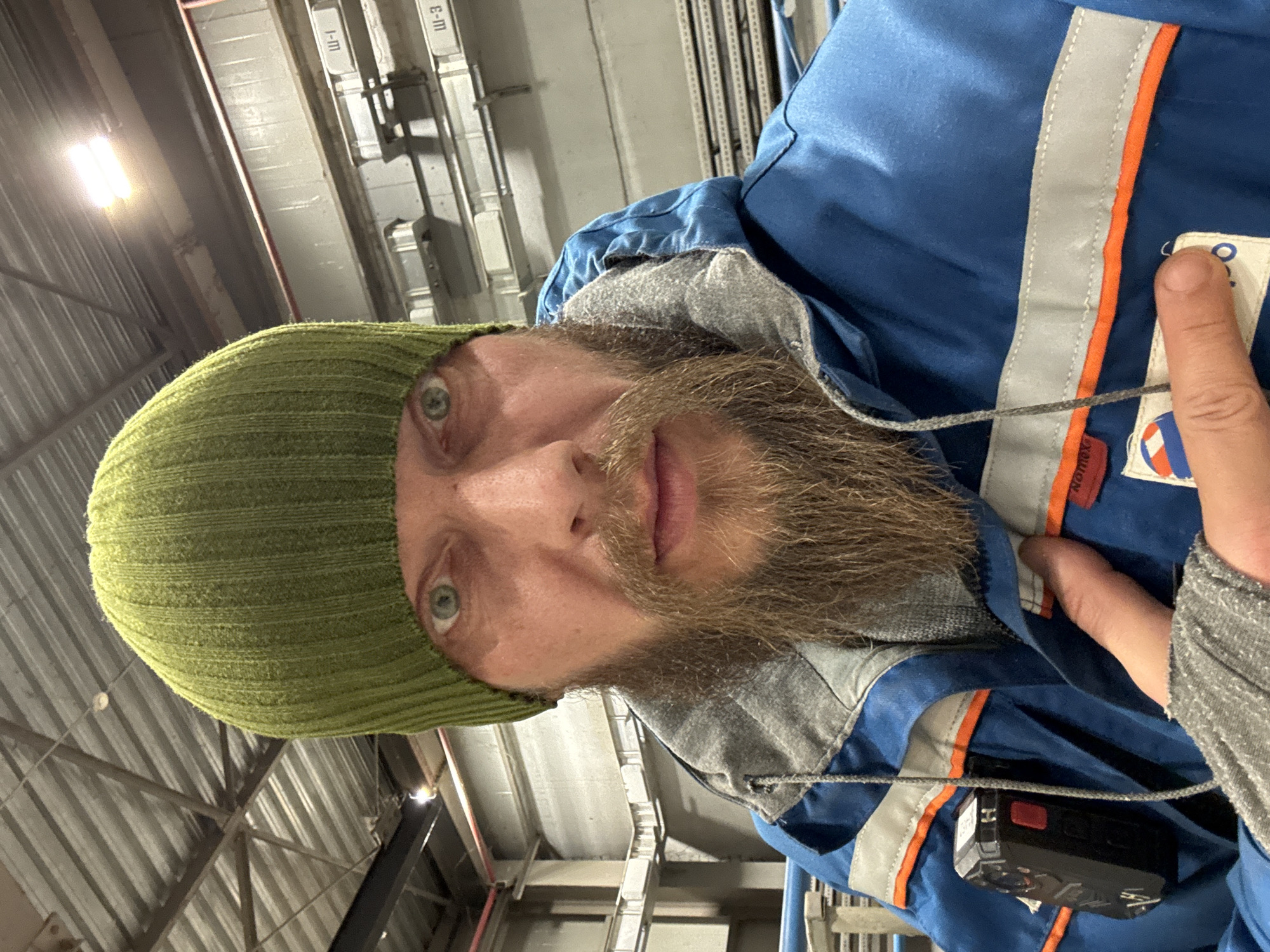 A man with a beard and green beanie is indoors. He is wearing a blue and grey jacket with reflective stripes. The background shows an industrial interior with metal structures and overhead lighting. The location in the city of Volzhsky, Russia cannot be confirmed from the image.