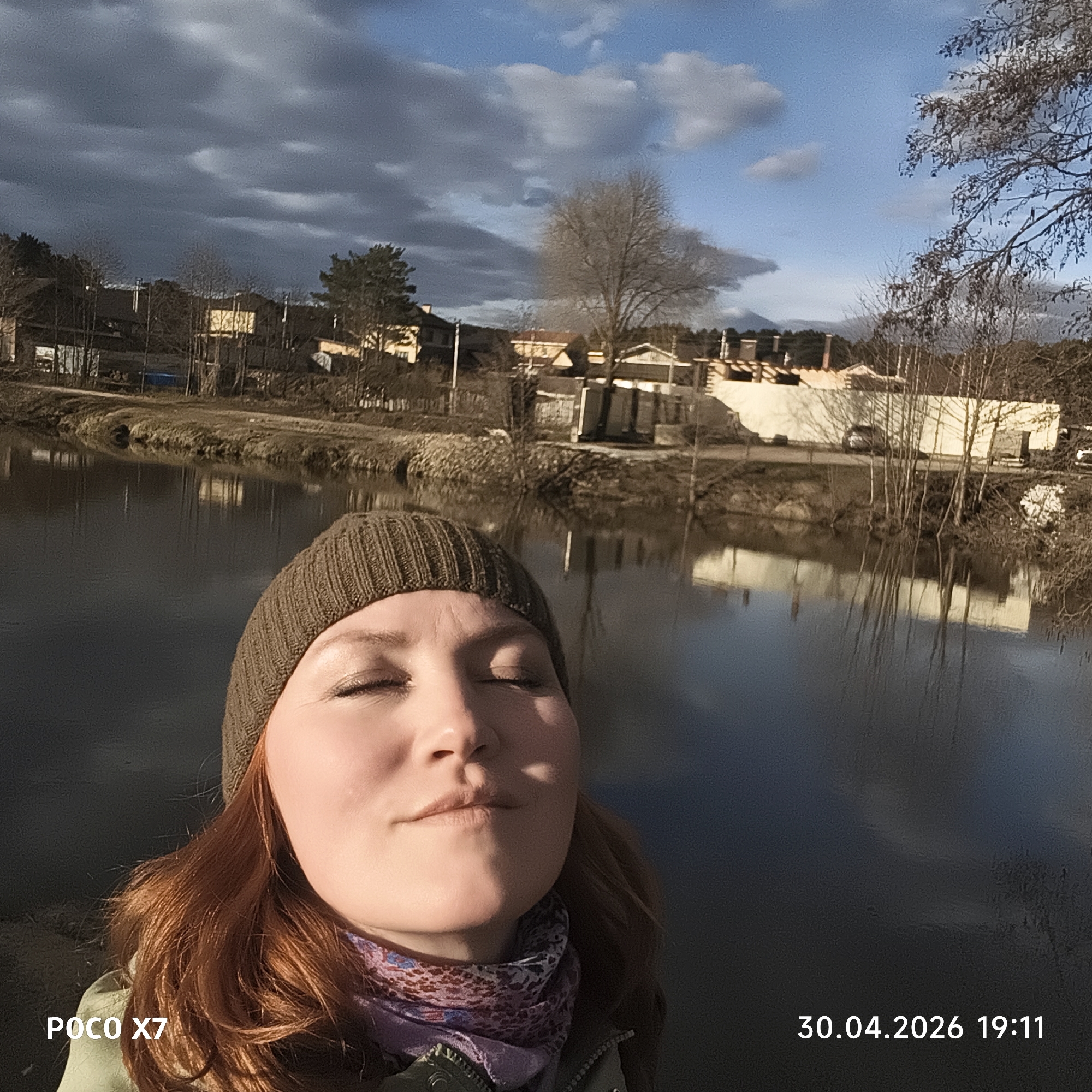 A woman with reddish-brown hair, wearing a brown knitted beanie and a patterned purple and white scarf, takes a selfie outdoors. Her eyes are closed, and her face is illuminated by strong sunlight, creating shadows and conveying a sense of peace and enjoyment.
Behind her is a wide body of water, likely a river or lake, reflecting the distant shoreline. The opposite bank features a developed landscape with various buildings, including houses and other structures, nestled among a mix of bare deciduous trees and evergreen pine trees. The trees suggest an early spring setting, as many are still without leaves. The buildings appear to be a combination of residential and possibly light industrial or commercial.
The sky is partly cloudy, with patches of bright blue visible between darker grey clouds. The strong, warm sunlight on the woman's face and across the scene, combined with the timestamp, indicates late afternoon or early evening. The scene is set in Chaikovsky, Russia.
Visible text in the bottom left corner states "POCO X7", and in the bottom right corner, the date and time "30.04.2026 19:11" are displayed.