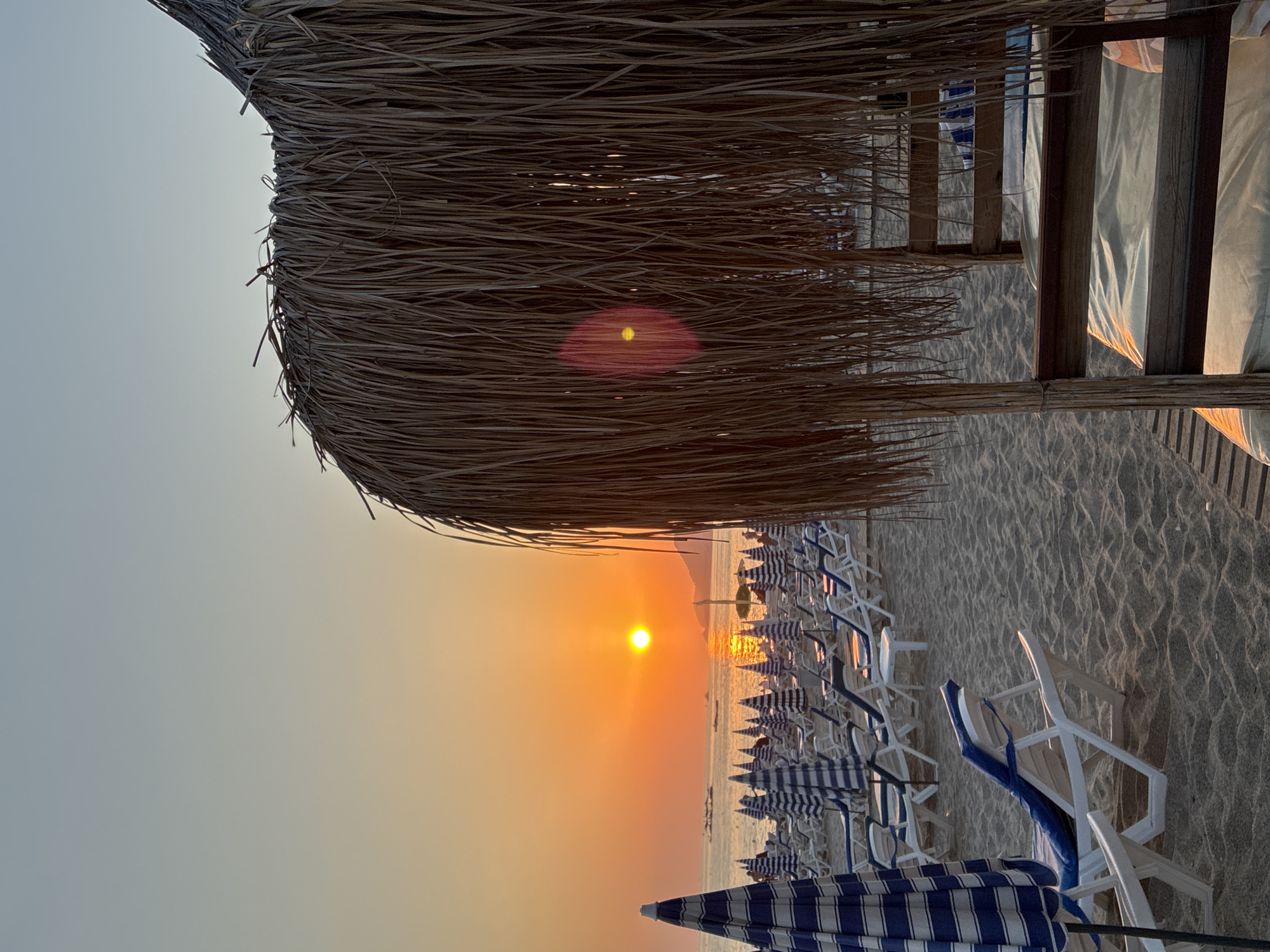 The image depicts a beach scene at sunset in Alanya, Turkey. The foreground is dominated by a thatched roof structure casting a shadow over a wooden lounge bed. To the left, a striped beach umbrella stands prominently. Rows of empty lounge chairs and umbrellas stretch across the sandy beach towards the sea. The sun, a bright orange orb, is setting over the calm water, casting a warm glow and its reflection on the sea. The sky transitions from a pale blue at the top to a vibrant orange near the horizon. Several boats are visible in the distance on the water. The overall impression is one of tranquility and a peaceful end to the day.