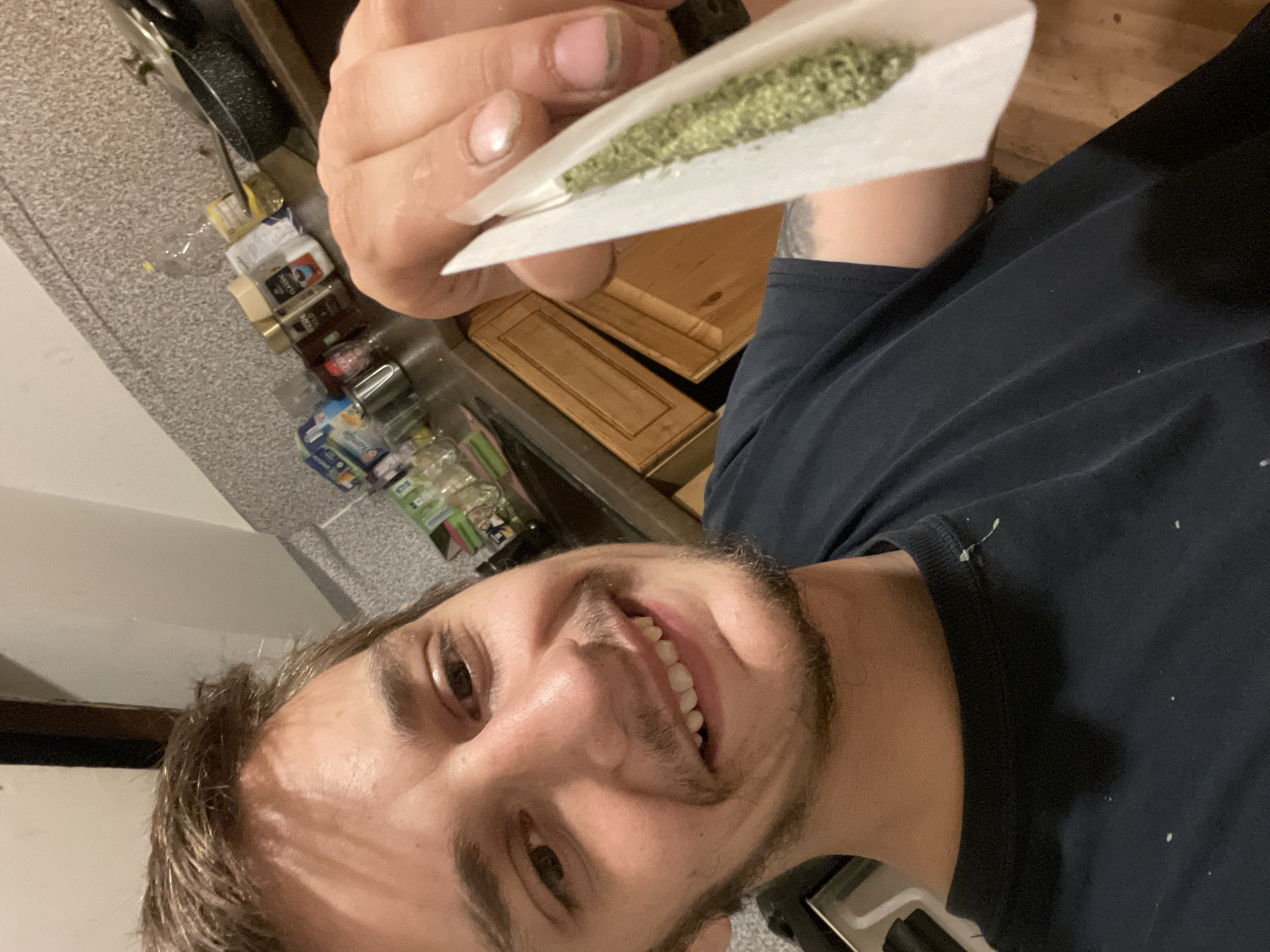 A selfie of a man with brown hair and a short beard, wearing a navy blue t-shirt, holding a partially rolled cigarette filled with green plant material. The background shows a kitchen counter with various items, including spice jars, canned goods, and cleaning supplies. Behind the counter are wooden cabinets.