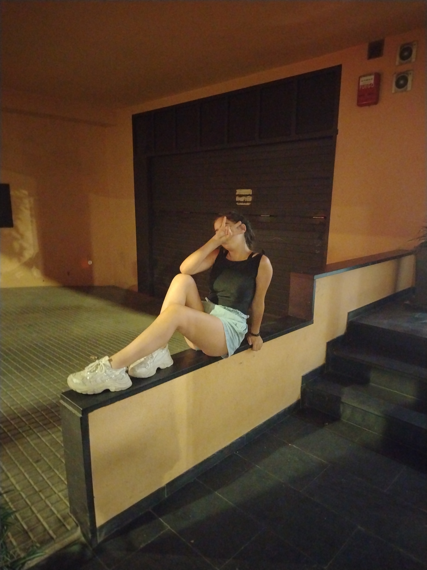 A young woman with dark hair, wearing a black tank top and light blue shorts, sits on a raised ledge with her legs extended. She is covering her face with her right hand and her left hand is near her face, possibly signaling a peace sign. She is wearing white sneakers. The ledge she sits on is part of a structure with a yellow-beige textured surface and a black border. Behind her is a dark, closed door with vertical paneling. To the right of the ledge, a dark staircase descends. The surrounding environment appears to be an outdoor area at night, with orange-toned lighting. Two fire alarm boxes are visible on the orange wall above and to the right of the door. The ground in front of the ledge is paved with grey tiles. The image was taken in Arbúcies, Spain.