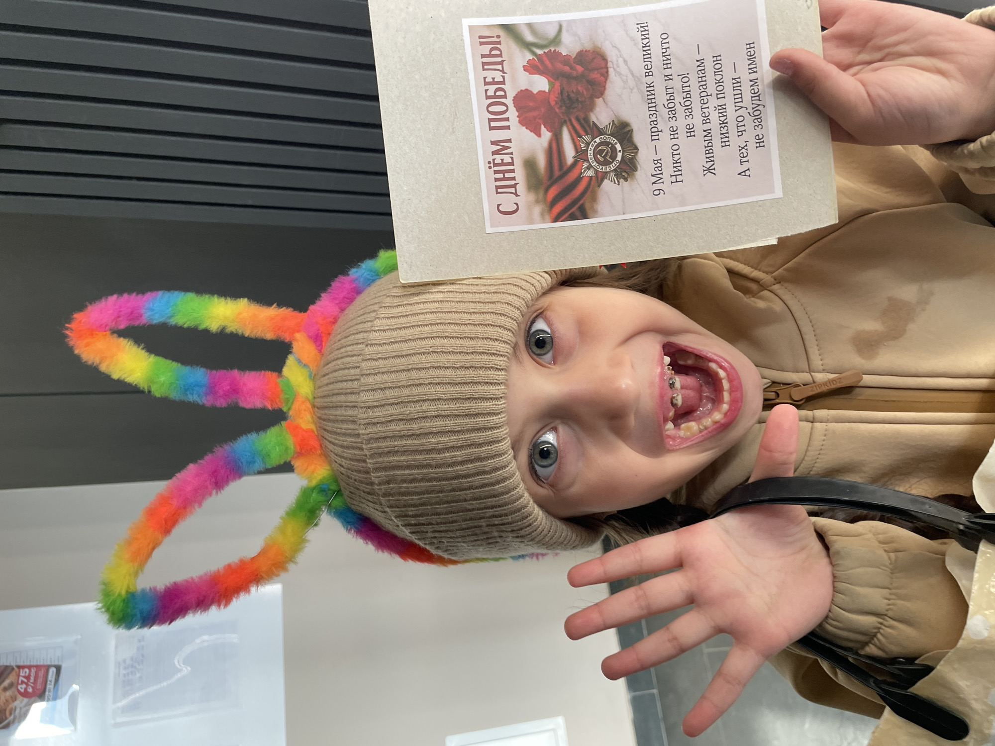 A young child wearing a beige knit hat with rainbow fuzzy antennae is upside down, holding a card that reads "Happy Victory Day!" in Russian. The card features an image of the Order of the Patriotic War medal, associated with World War II. The child's face shows a wide, toothy grin, revealing dental braces. They are wearing a tan jacket and holding a black strap. In the background, there is a ceiling with horizontal slats and, in the bottom left corner, what appears to be a price tag with the number 475 on it. Part of the Russian city of Izhevsk is visible as well.