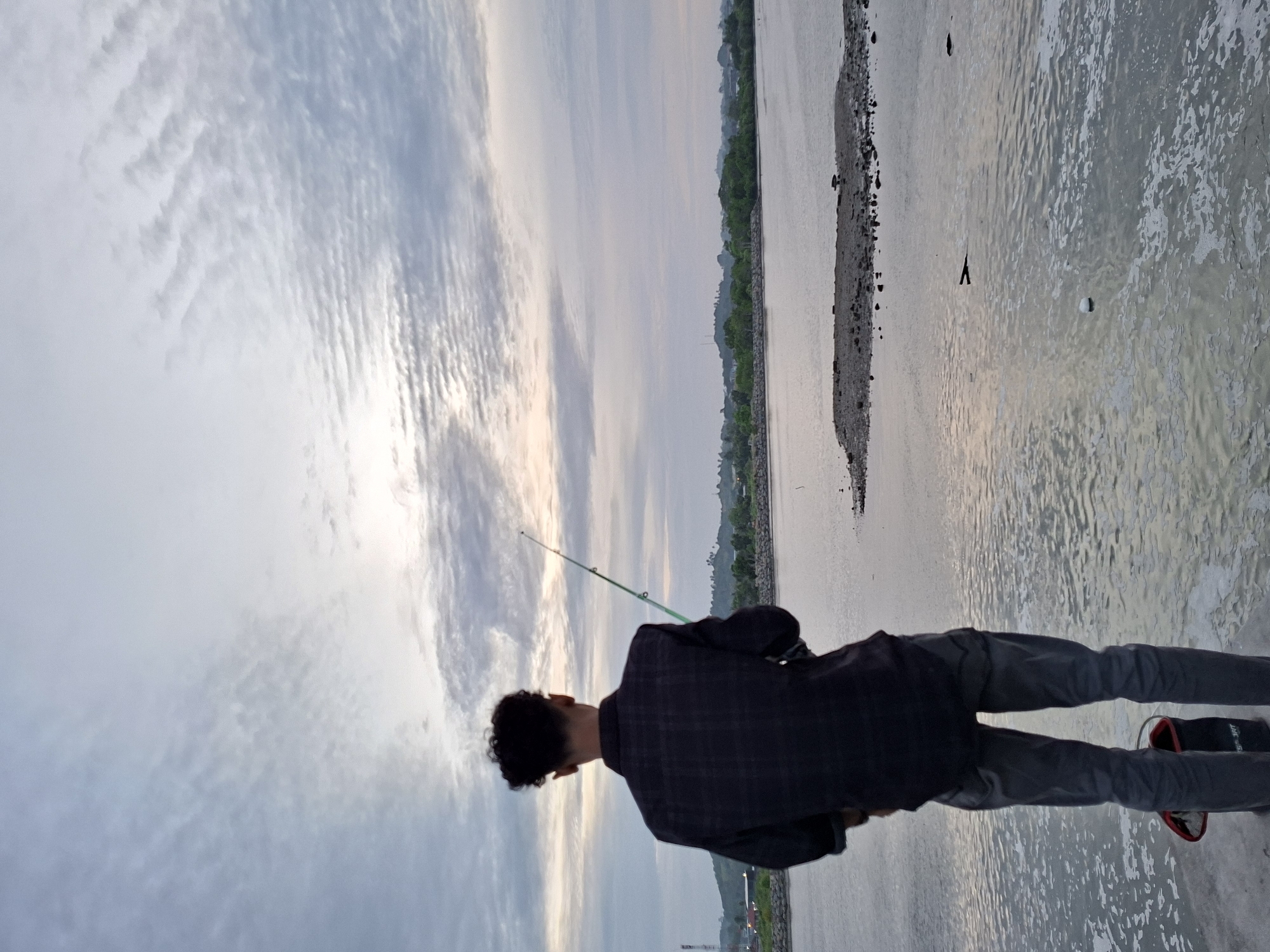 A man in a plaid jacket and gray pants stands on a concrete structure beside water, holding a fishing rod, with the sky above filled with clouds. The body of water is next to a strip of land with green trees. The location is in Lhokseumawe, Indonesia.