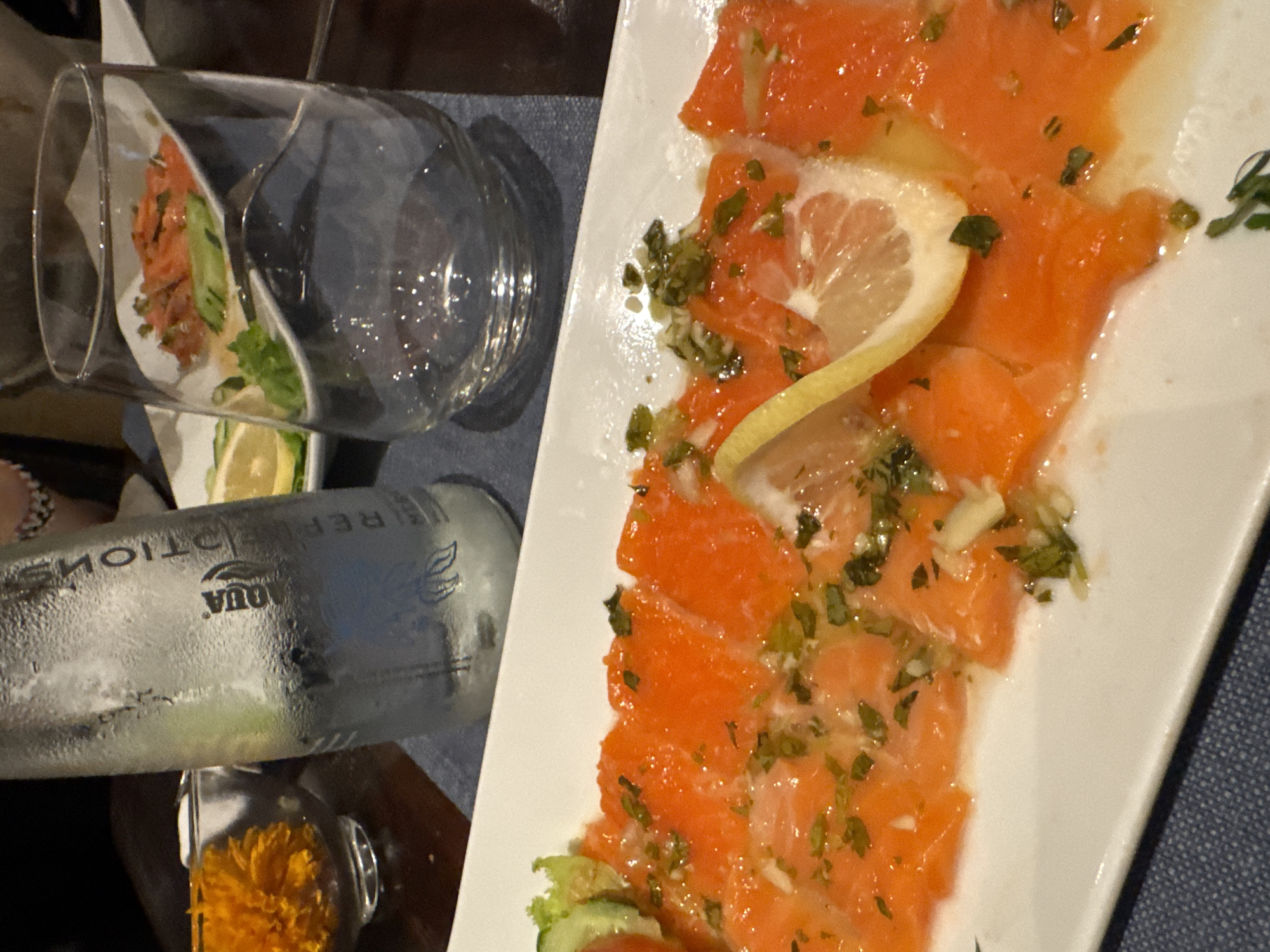 A close-up, overhead shot depicts a dish of thinly sliced salmon carpaccio arranged on a white rectangular plate. The salmon is bright orange and appears to be dressed with a light vinaigrette, chopped herbs, and thinly sliced garlic. A lemon slice rests on top of the carpaccio, and some lettuce and cucumber are visible on the left side of the plate. To the left of the plate, a condensation-covered bottle of Aqua brand mineral water stands upright. Behind the salmon, a clear glass rests at an angle, and a small white plate with more salmon and greens is visible through it. In the background, a small orange flower in a glass bowl and a blurry figure wearing a patterned bracelet are present. The setting appears to be a restaurant table in North Kuta, Indonesia, with a dark wooden surface and a dark blue placemat underneath the plate and water bottle.