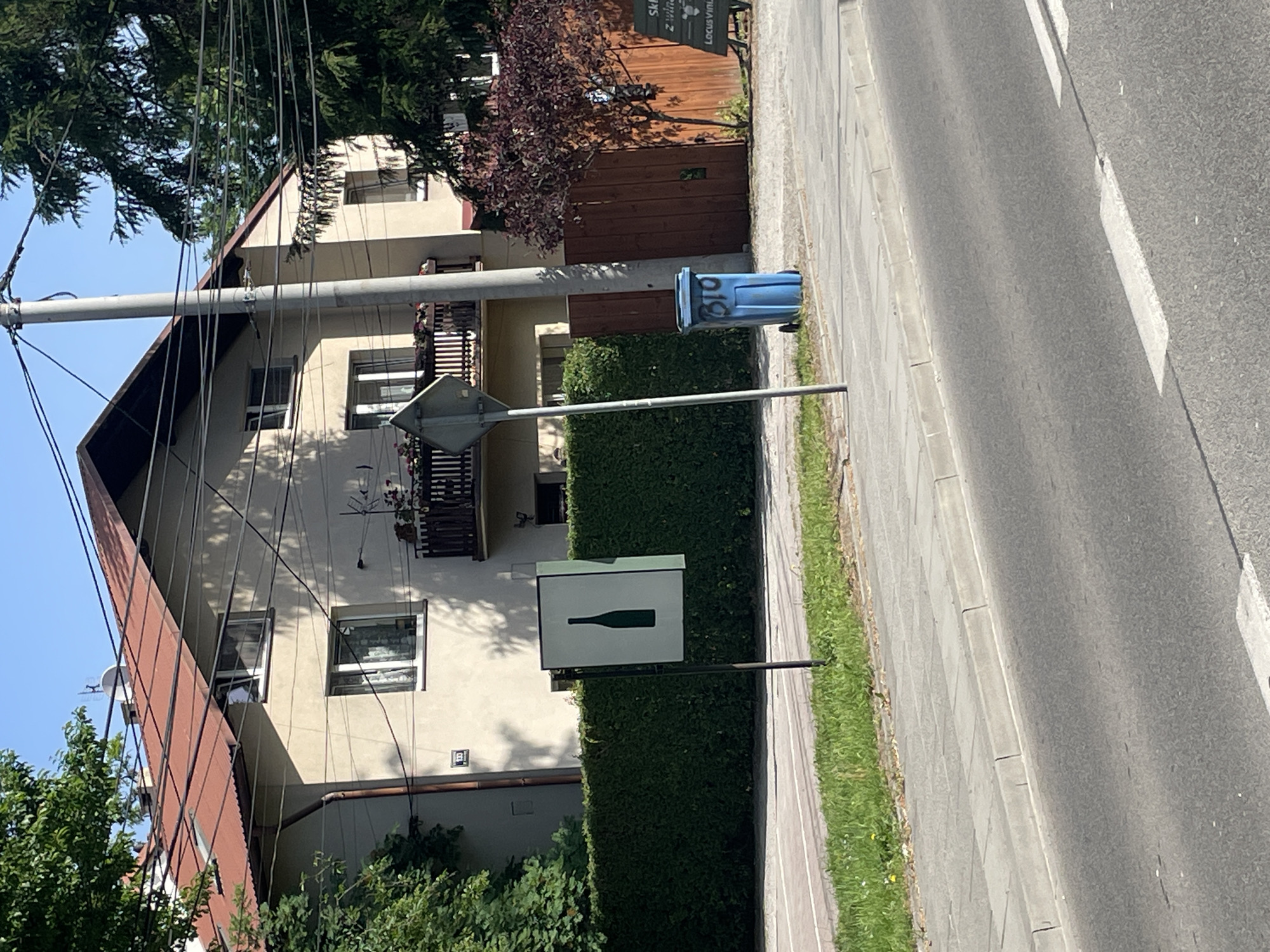 The image shows a street scene in an unidentified city. A multi-story building stands to the left, featuring windows, balconies, and a pitched roof. Overhead power lines are visible. A blue trash can sits on the sidewalk. A street sign with a bottle symbol is partially obscured by a hedge. The street stretches out along the right side of the image.