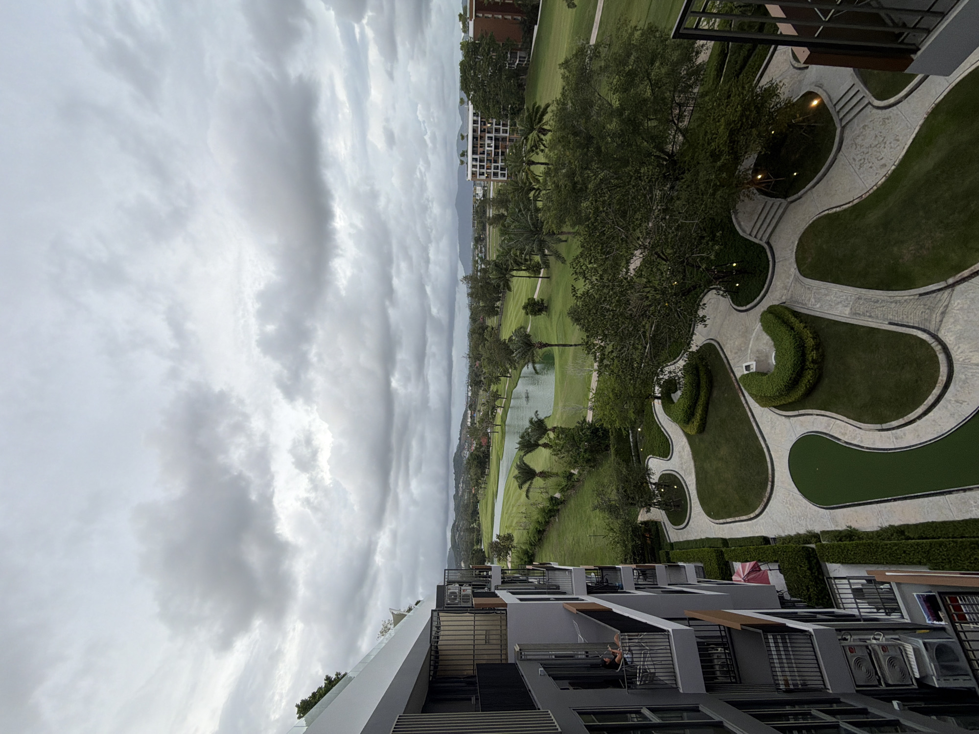 An elevated view, likely from a balcony in a building, reveals a scene in Choeng Thale, Thailand. Below, manicured green spaces and walkways define the landscape, interspersed with trees, a pond, and buildings. In the distance, other structures are visible, appearing to merge into the horizon. The foreground showcases the architecture of the buildings from which the photo is taken, including balconies and exterior units. Above, a heavy sky filled with grey clouds dominates the upper portion of the image, suggesting overcast weather.