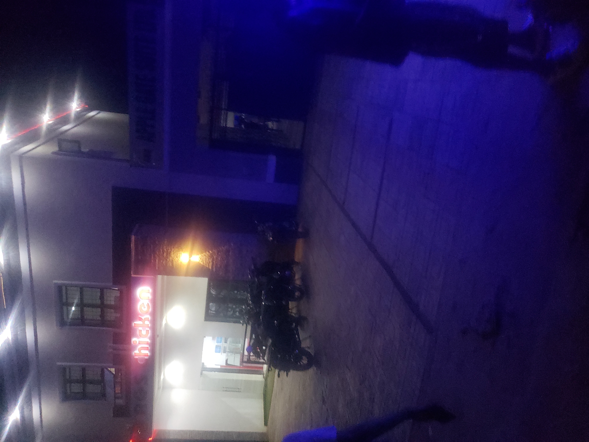 This image captures an outdoor commercial scene at night, located in Rido, Kaduna, Nigeria, specifically featuring a part of the Apple Gate Suites and Event Centre complex.
The scene is dominated by a two-story building with a white facade. The left section of the building displays two dark-framed windows on the upper level. Below these windows, a red neon sign reads "hicken" (likely "Chicken"), indicating a restaurant or fast-food establishment. The entrance beneath this sign is brightly illuminated, revealing a white interior space. To the right of this entrance, a textured dark wall section features a single orange-yellow wall sconce.
The right section of the building, connected to the left, is slightly set back and darker. Above a recessed archway or entrance, a sign is partially visible, reading "APPLE GATE SUITE," identifying the larger establishment. Linear red lights or reflections are visible on the upper facade of this section. The entrance itself is dark.
The ground in the foreground is a paved surface, likely concrete, with visible seams and variations in lighting. Three dark-colored motorcycles are parked directly in front of the "hicken" establishment. A fourth motorcycle is partially visible further to the right.
Two individuals are partially visible in the foreground. On the far left, the arm and upper torso of a person wearing a light blue garment are visible. On the far right, the lower body of a person, clothed in dark attire, is strongly illuminated by a pervasive blue light source originating from outside the frame on the right. This blue light casts a significant color tint across the right half of the paved area. Another person's legs are dimly visible further back within the blue-lit zone. Overhead light sources on the building provide ambient illumination, creating a high-contrast environment with both bright spots and deep shadows.