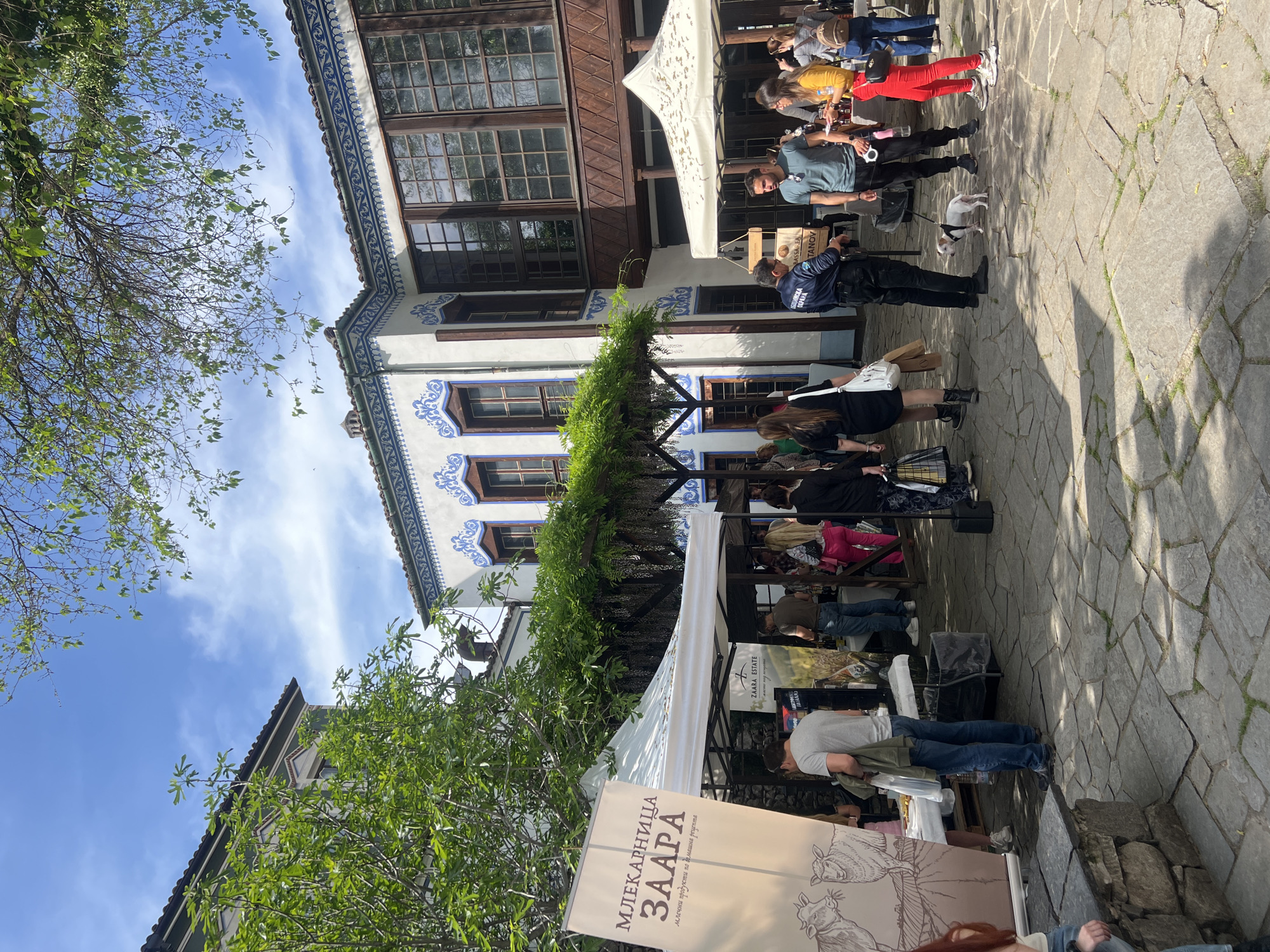 The image captures a scene in Plovdiv, Bulgaria, featuring an outdoor market or event. The distinctive Bulgarian National Revival architectural style building with white walls and blue ornamental patterns is visible in the background. People are browsing stalls or booths under white canopies, with vendors serving customers. A small dog is walking amongst the people. Signage in Bulgarian is seen at the front. The ground is paved with stone blocks. Green trees and a blue sky are overhead.