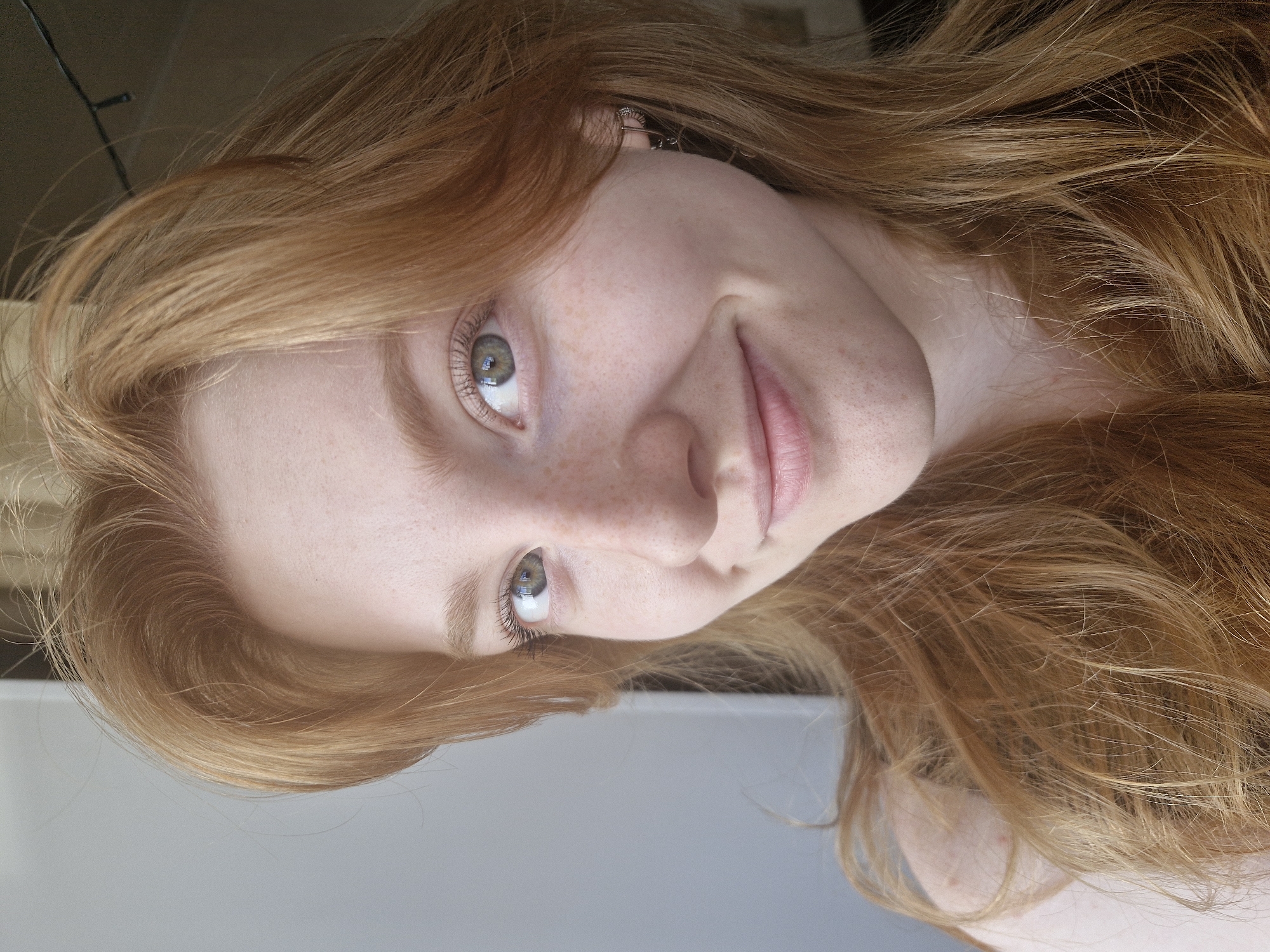 A close-up shot of a light-skinned person with red hair and freckles is shown. The person's face is tilted, so that the right eye is slightly higher in the frame. The person is smiling slightly. A small piece of jewelry is visible on the person's left ear. The background is blurred and features a white or gray object. There is no indication the photograph was taken in the city of Yekaterinburg, Russia.