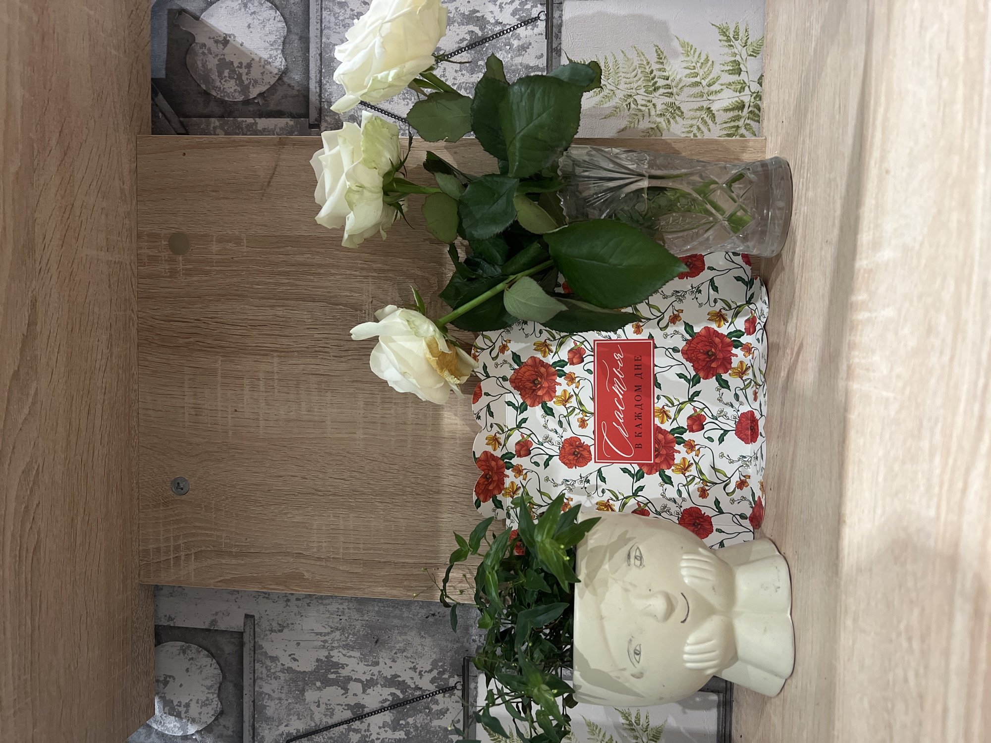 A still-life arrangement is positioned on a light brown, wood-grain surface, potentially the interior shelf of a cabinet or other furniture. The composition includes three white roses, several of which are in a glass vase, resting atop a decorative box adorned with red flower illustrations and a red label with Cyrillic script. Adjacent to the box is a white ceramic head planter filled with green leafy foliage. The backdrop consists of two distinct wallpapers. One wallpaper exhibits a gray, mottled pattern, while the other features a print of green ferns. The scene is indoors. There is no explicit indication of Sochi, Russia.