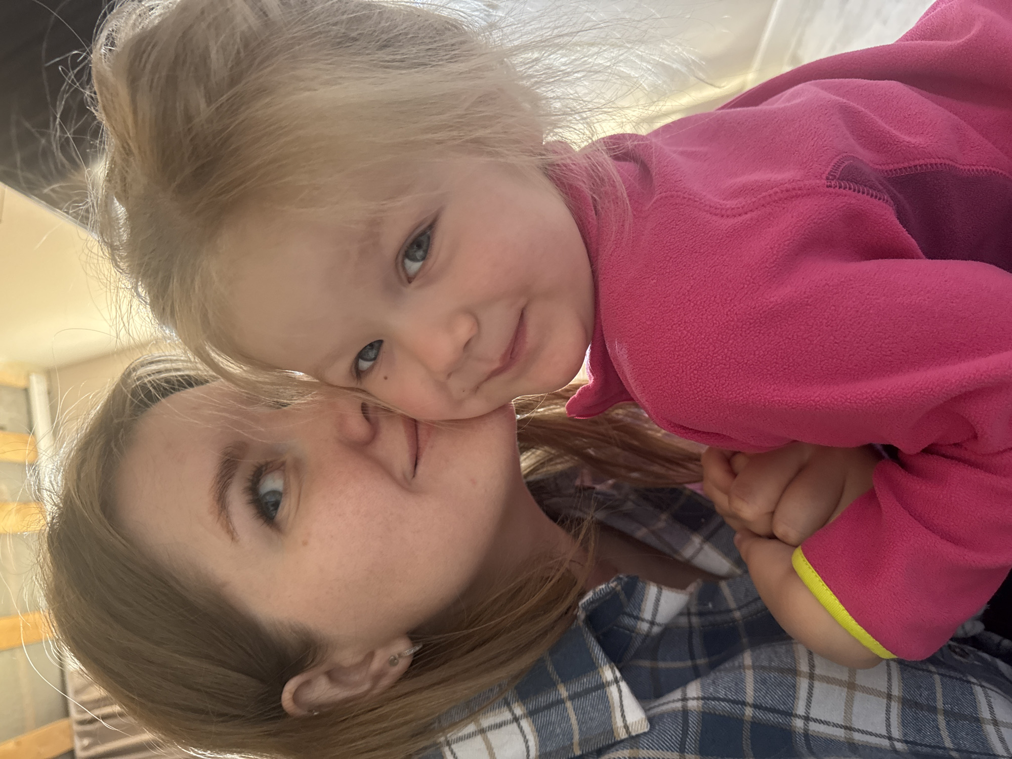 A young child with blonde hair and blue eyes, wearing a pink fleece jacket with a yellow trim on the sleeve, is being held by a woman with light brown hair, blue eyes, and freckles, who is wearing a blue and white plaid shirt. The child's face is close to the woman's face, suggesting affection. The setting appears to be indoors, with a glimpse of a window frame in the background. There is no indication of the location being in Gornyi (Dvurechenskoe S/P), Russia within the image.