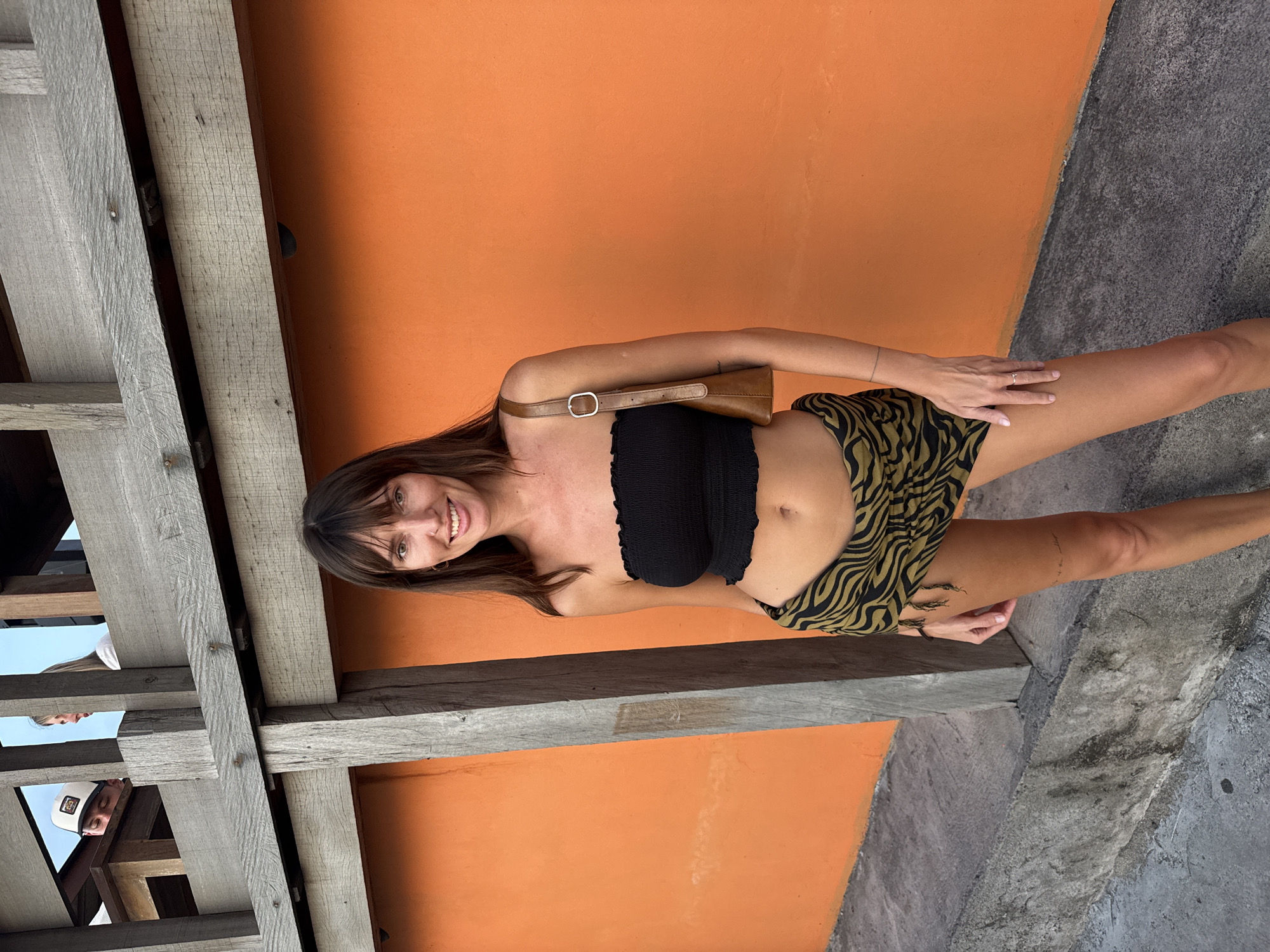 The image shows a woman standing in front of an orange wall, seemingly in North Kuta, Indonesia, based on the architectural details and general ambiance. She is wearing a black strapless top and a patterned skirt. She carries a brown bag with a strap over her shoulder. Wooden beams are visible overhead. The flooring is concrete. Several blurry faces are seen on the left. The scene is well-lit, seemingly taken outdoors.