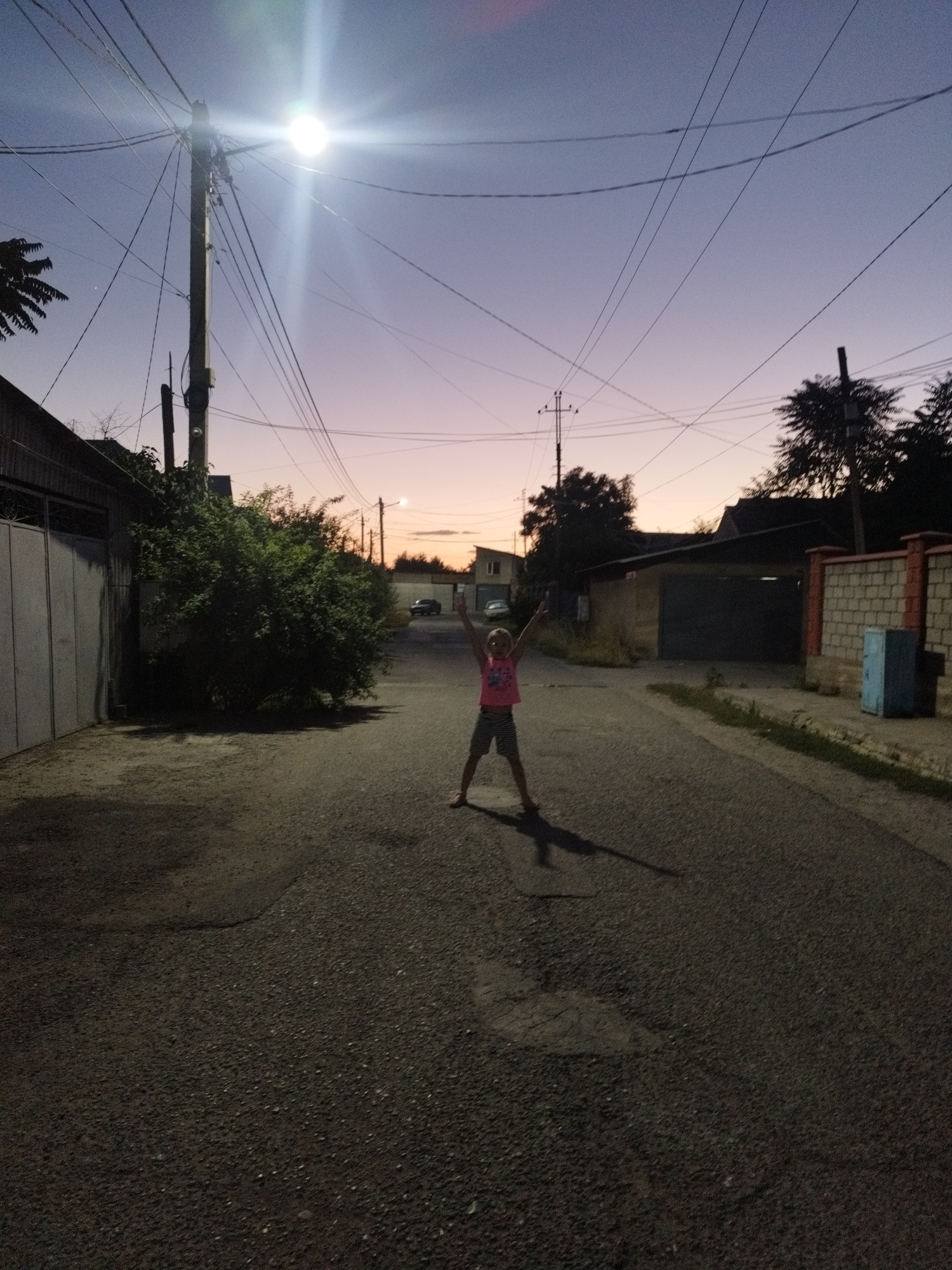 In this nighttime photograph, a young person stands in the middle of a paved street, arms raised in celebration. The sky above transitions from a deep twilight blue to a soft lavender and then a pale peach near the horizon, indicating it is either dusk or dawn. A bright streetlight illuminates the scene, casting long shadows.
The setting appears to be a residential street in Shymkent, Kazakhstan, with houses and fences lining the sides of the road. To the left, a large grey garage door is visible, adjacent to a dense green bush. On the right, a brick fence encloses a property, with a light blue cabinet or utility box next to it. In the distance, a car is parked on the street, and trees are silhouetted against the colorful sky.
The person in the foreground is wearing a pink top and striped shorts, their stance suggesting joy or excitement. The overall mood of the image is peaceful and a little magical, capturing a moment of freedom and youthful exuberance under the soft glow of the streetlight and the fading sky. The ground is asphalt, with some patches and cracks visible, common in urban or suburban environments. No visible text is present in the image.