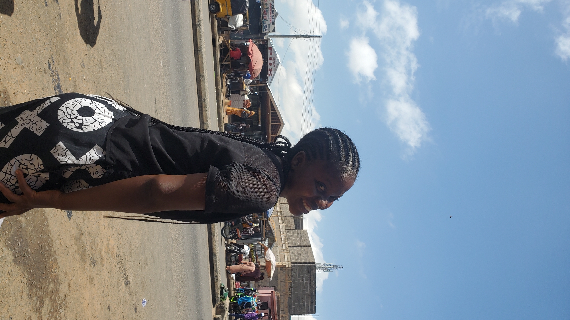 A young woman with braided hair is standing in an outdoor market on a sunny day. She is wearing a black top and black and white patterned pants. She is looking up and to her right, with a smile. In the background, there are market stalls with vendors, people shopping, and motorcycles. The sky is blue with scattered white clouds.