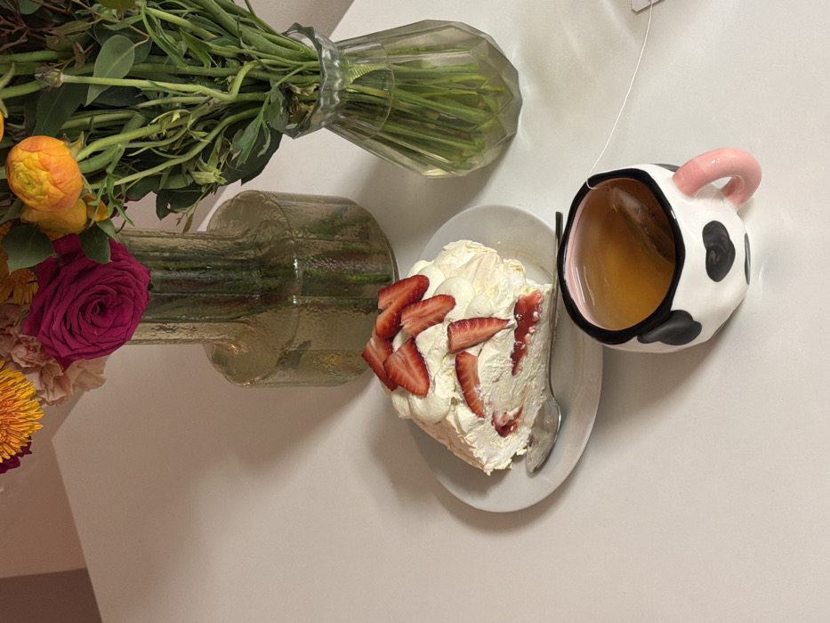 The image depicts an overhead view of a white table surface in Tel-Aviv, Israel, under direct illumination casting soft shadows. A slice of strawberry meringue roulade, featuring whipped cream and fresh strawberry slices atop a meringue base with strawberry filling, rests on a white circular plate. A silver spoon is positioned next to the dessert. To the right, a white ceramic mug with irregular black spots, mimicking a cow print, and a pink handle, contains an amber-colored liquid with a white tea bag string visible. Behind these items, two vases hold floral arrangements. The larger, amber-tinted, vertically ribbed glass vase contains a bouquet including a prominent fuchsia rose, several yellow and orange ranunculus-type flowers, and green foliage. The smaller, clear, vertically faceted glass vase contains additional green stems and foliage, with some yellow-orange flower tips visible. No individuals are present in the scene.