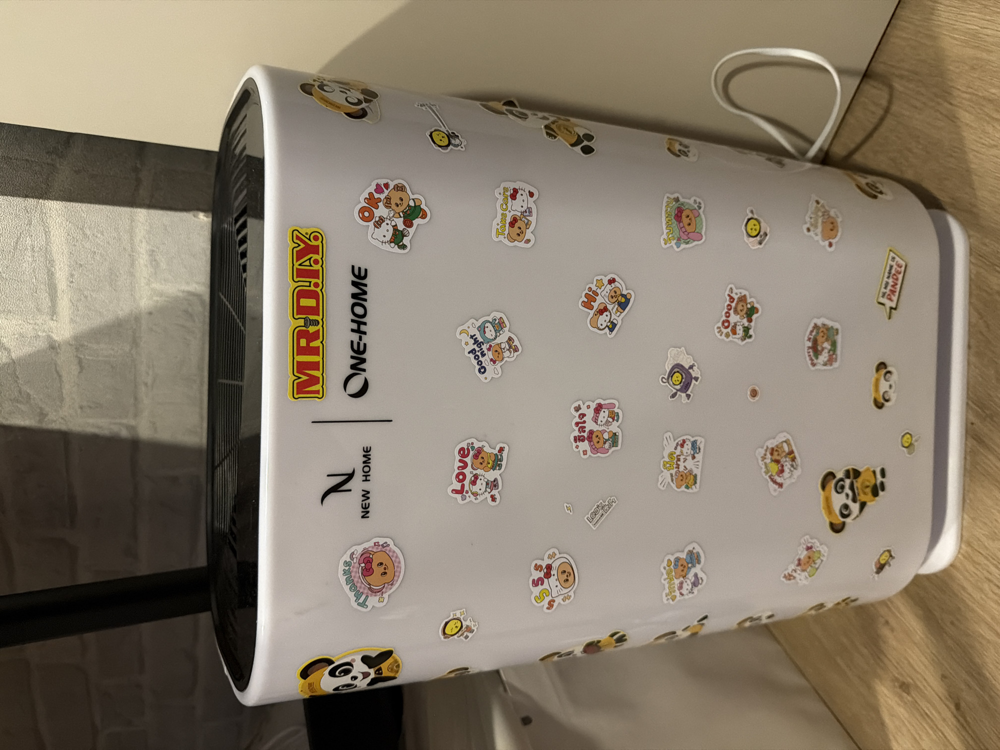 The image shows the side of a white air purifier with several colorful stickers adhered to its surface. The air purifier is placed on a wooden floor, with a textured brick-patterned wall in the background. A black cable is visible on the right side of the air purifier, extending downwards. The stickers depict cartoon characters, including pandas, bears, and other animals, with various phrases like "Thanks", "Love", "OK", "Good night", "Hi", and "Take Care". There are also some stickers with text in Thai script. The overall scene appears to be an indoor domestic setting, likely a home or office. The air purifier itself is a modern appliance, suggesting it is a functional item within the environment. There are no people or animals present in the image. The lighting suggests a moderately lit indoor environment.