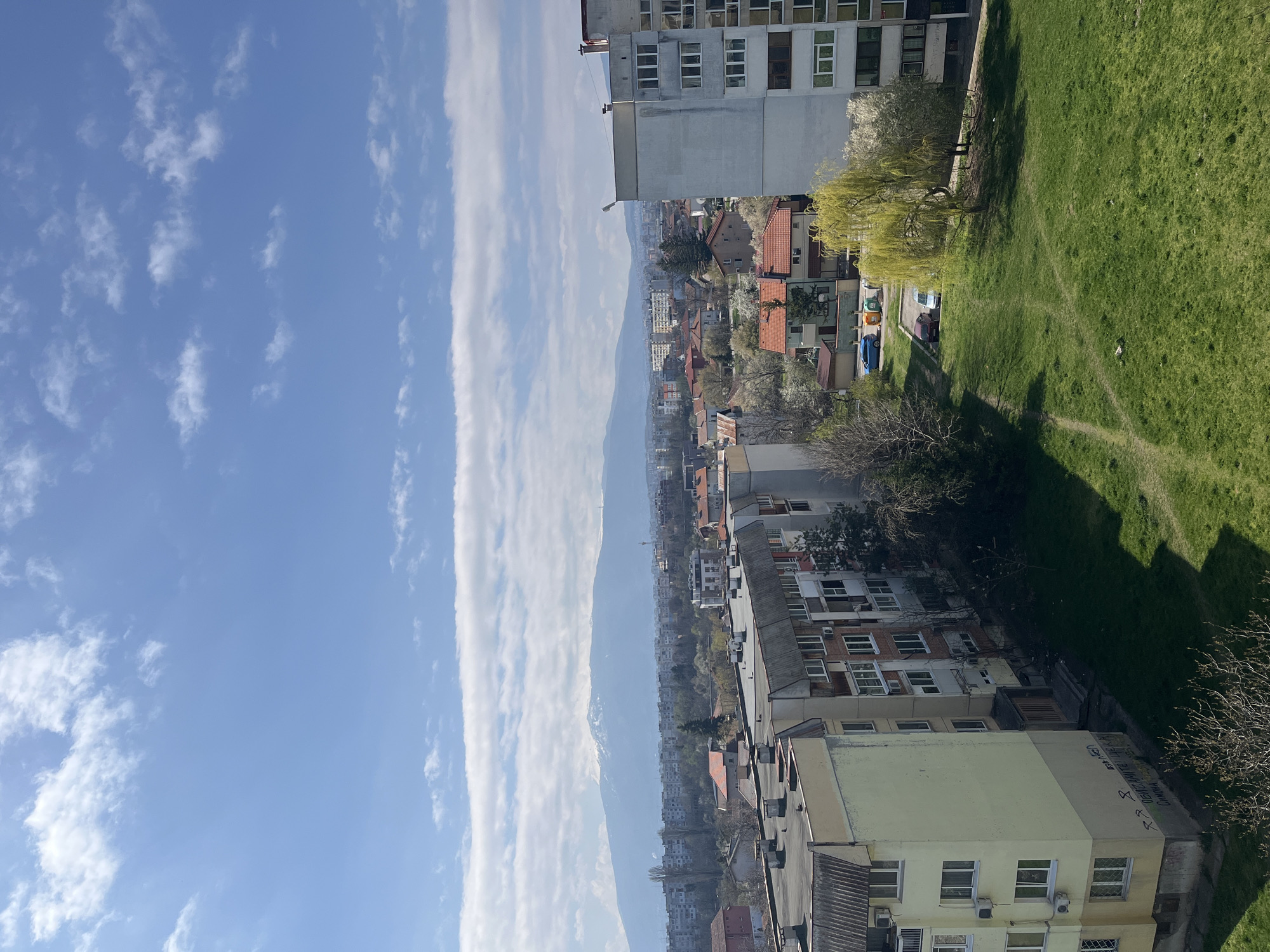 The image depicts a cityscape in Sofia, Bulgaria. A wide, partly cloudy blue sky occupies the left portion of the frame, contrasted by rows of buildings and residential areas on the right. The buildings are a mix of apartment blocks and houses with red tile roofs. A long, elongated mountain range is visible in the distance. A patch of green grass with sparse trees is located on the right side of the image near the top corner. There are no people visible in the image.