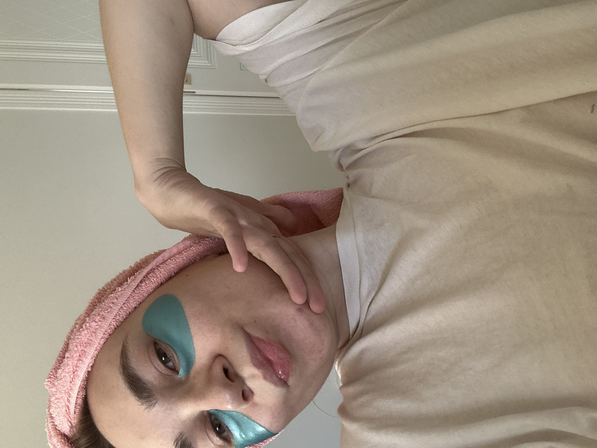 An upward-looking, close-up shot of a person, rotated 90 degrees clockwise, captures them engaged in a self-care routine in what appears to be an indoor setting in Kharnavy, Russia. The person has fair skin, prominent dark eyebrows, and dark eyes. Two teal-colored, teardrop-shaped under-eye patches are applied beneath their eyes. A pink terry cloth towel is wrapped around their head, suggesting post-shower or hair treatment. They are wearing a light, possibly beige or off-white, top. One of their hands is visible, with fingers gently resting near their cheek. Their lips are glossy, and a small reddish blemish is visible on their cheek. The person's expression is neutral, looking directly towards the viewer or camera. In the background, a light-colored wall or door with simple horizontal molding is visible, reinforcing the indoor, private nature of the scene. No specific time of day or weather is discernible, and there is no visible text.