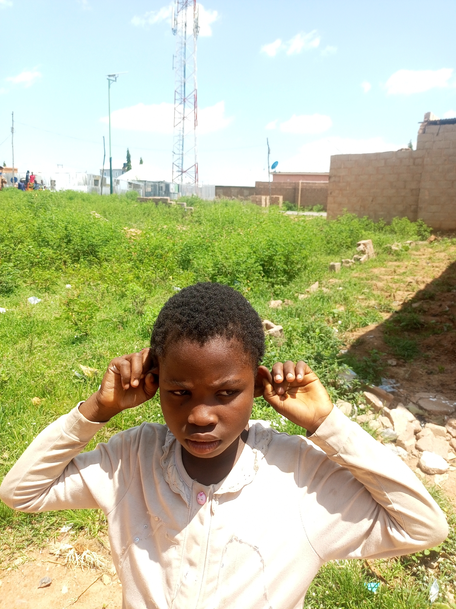 A young Black girl is shown in the foreground, her hands covering her ears. She wears a light-colored long-sleeved shirt with a ruffled collar and a pink button. Her expression is serious as she looks slightly off-camera. Behind her, a grassy field is visible, with scattered debris and rocks. In the middle ground, a tall telecommunications tower stands prominently against a bright, cloudy sky. Various buildings and structures, some appearing incomplete, are scattered in the background. The environment suggests an outdoor setting in a developing urban or semi-urban area, possibly Tempure, Nigeria, characterized by a mix of industrial infrastructure and unpaved land.