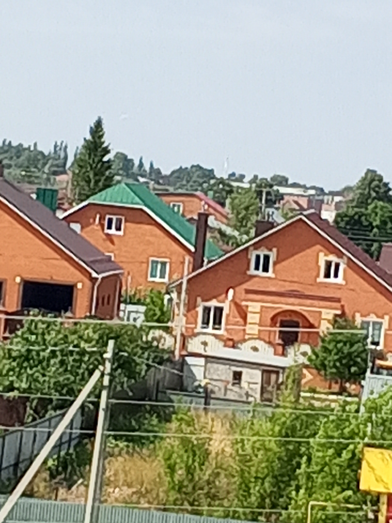 The image depicts a residential area with several brick houses, likely in the city of Chistopol, Russia, given the context. The houses are varied in design, with some featuring prominent gables and green or brown tiled roofs. Windows are visible on the houses, some with white frames. A large tree stands in the background, and dense greenery, possibly gardens or trees, surrounds the houses. A fence is visible in the foreground, with some structures partially obscured by vegetation. The sky is a pale, overcast grey. The scene appears to be a sunny day.