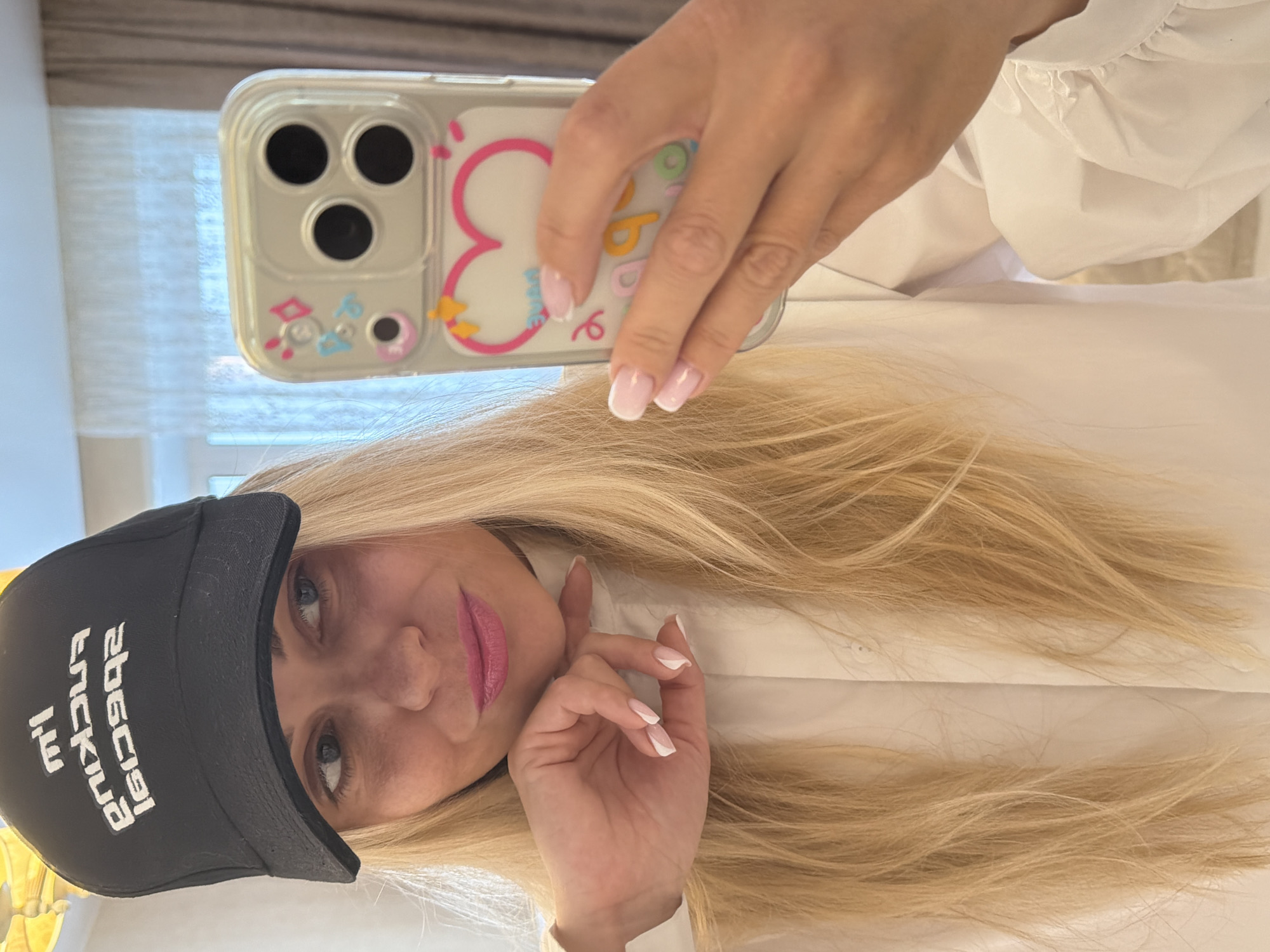 A woman is taking a selfie, holding a phone in her right hand. The phone has a clear case with pink and yellow embellishments and a visible triple-lens camera system. The woman has long blonde hair and wears a black baseball cap with white lettering. She is wearing pink lipstick and has manicured nails with white tips. She wears a white shirt or blouse. The background includes a window with closed blinds. There is no evidence to suggest a location in Votkinsk, Russia.