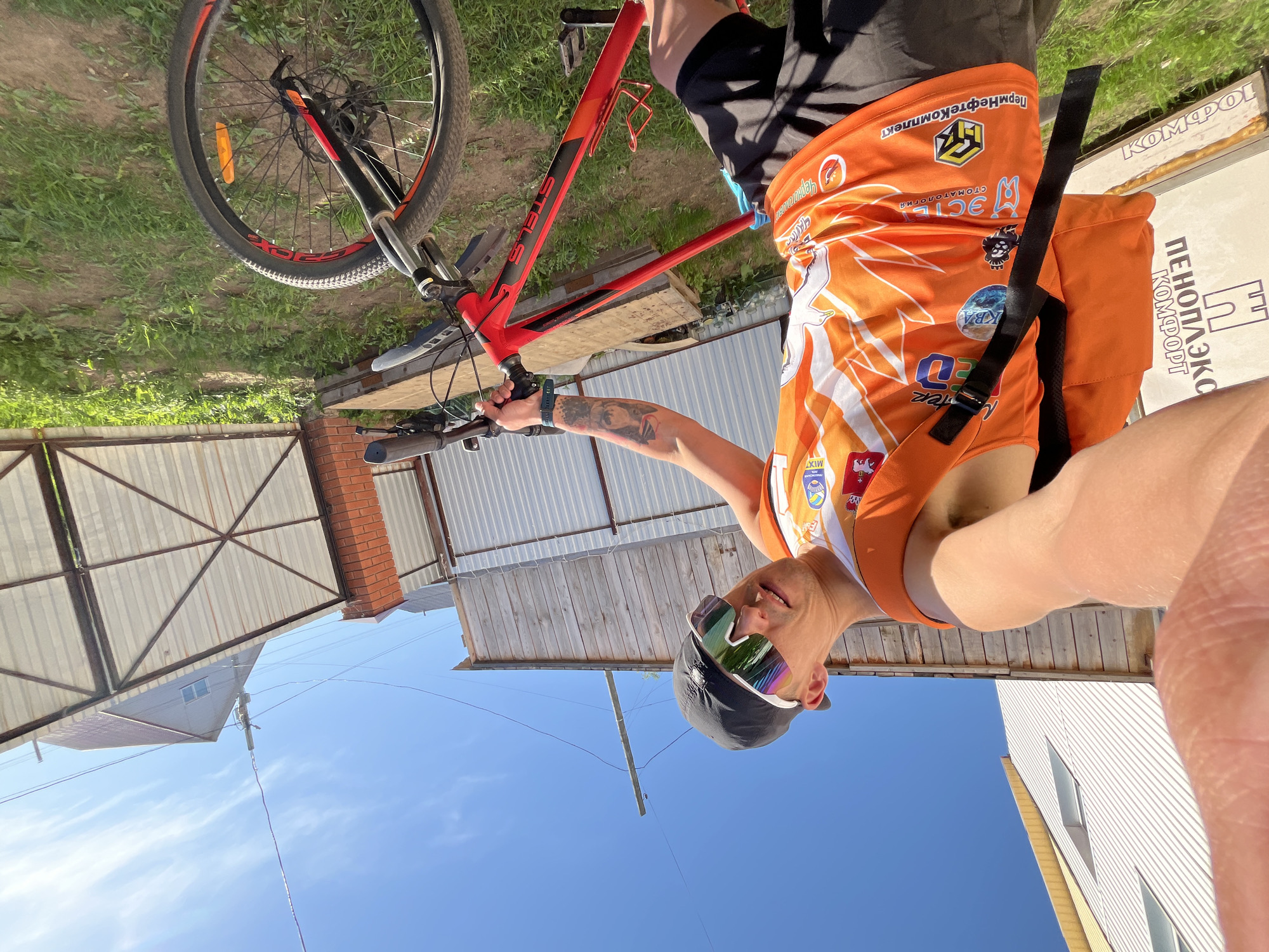 A person in a bright orange jersey and a black cap, wearing sunglasses, is taking a selfie while holding a red bicycle. The person has a tattoo on their left arm. The background shows a clear blue sky and a residential area with houses and fences. The sign "ПЕНОПЛЭКС" can be seen in the background. The location is likely a residential area within the city of Chaikovsky, Russia.