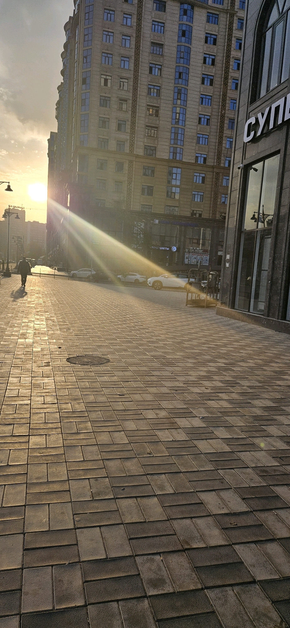 The image depicts a street scene in Dushanbe, Tajikistan, bathed in the golden light of a setting sun. The foreground is dominated by a paved walkway made of rectangular interlocking bricks, laid in a herringbone pattern. A man walks away from the camera towards the left of the frame, casting a long shadow on the bricks. To the right of the walkway, a modern building with dark facade and large glass windows is visible, with the Cyrillic word "СУПЕР" (SUPER) emblazoned on its exterior. Behind this, a taller, multi-story building with a beige facade and numerous blue-tinted windows rises into the sky. The sun is low on the horizon, casting strong rays of light and lens flare across the scene, illuminating the clouds and the buildings. Several cars are parked along the street to the left of the walkway, including a white sedan. A street lamp stands on a pole near the sidewalk. The overall atmosphere is one of urban tranquility during sunset.