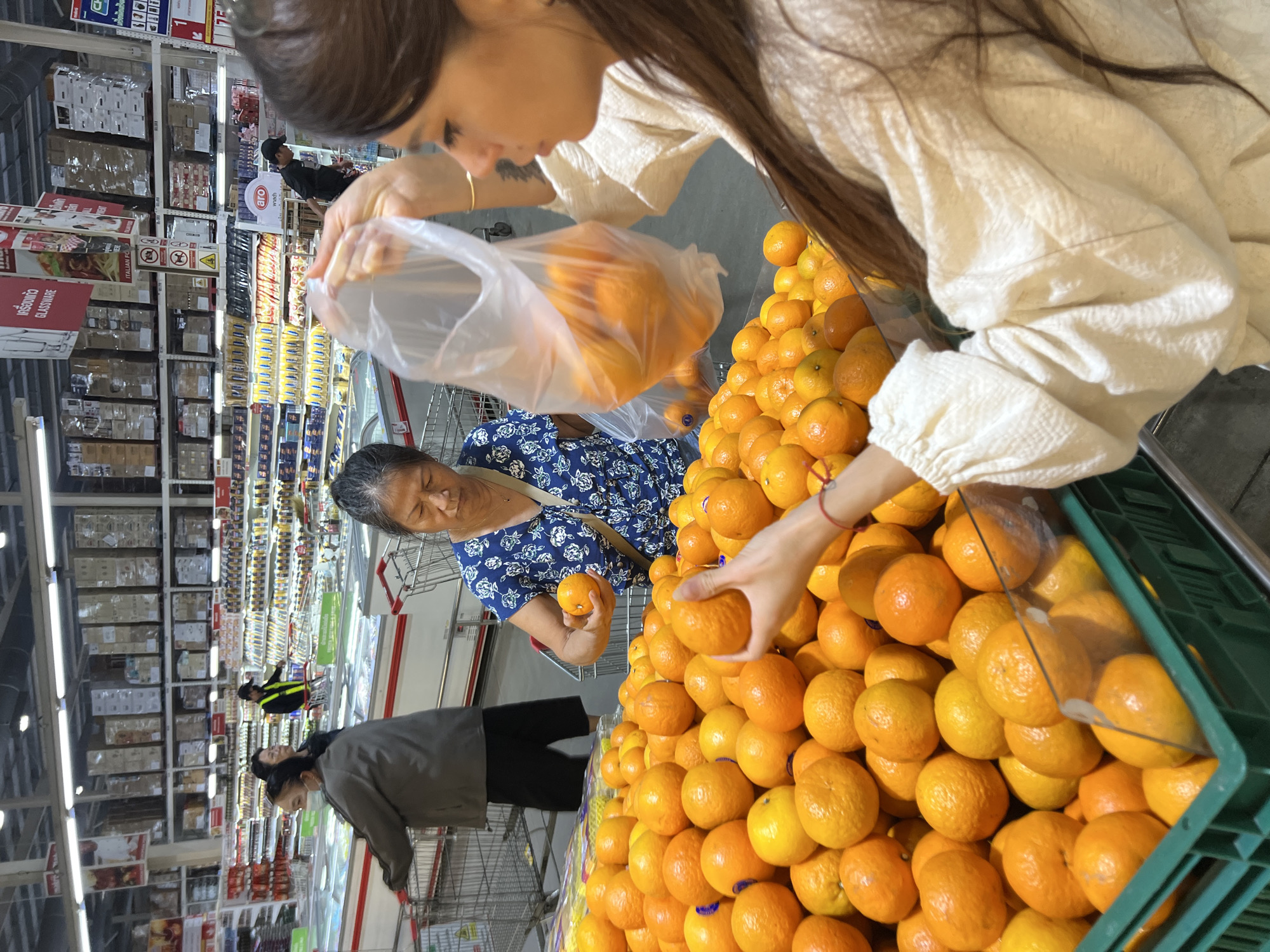 In Baan Bo Phut, Thailand, a grocery store scene unfolds with several individuals browsing produce. A woman in a blue floral shirt is selecting oranges, holding one in her hand, with a large display of oranges in the foreground. Another woman, on the right, is holding a clear plastic bag filled with oranges. In the background, a person wearing a mask and a grey jacket is visible, pushing a shopping cart. A man in a black cap and vest is also seen in the background, near shelves stocked with various goods. The environment appears to be a well-lit supermarket with aisles of refrigerated units and towering shelves of products.