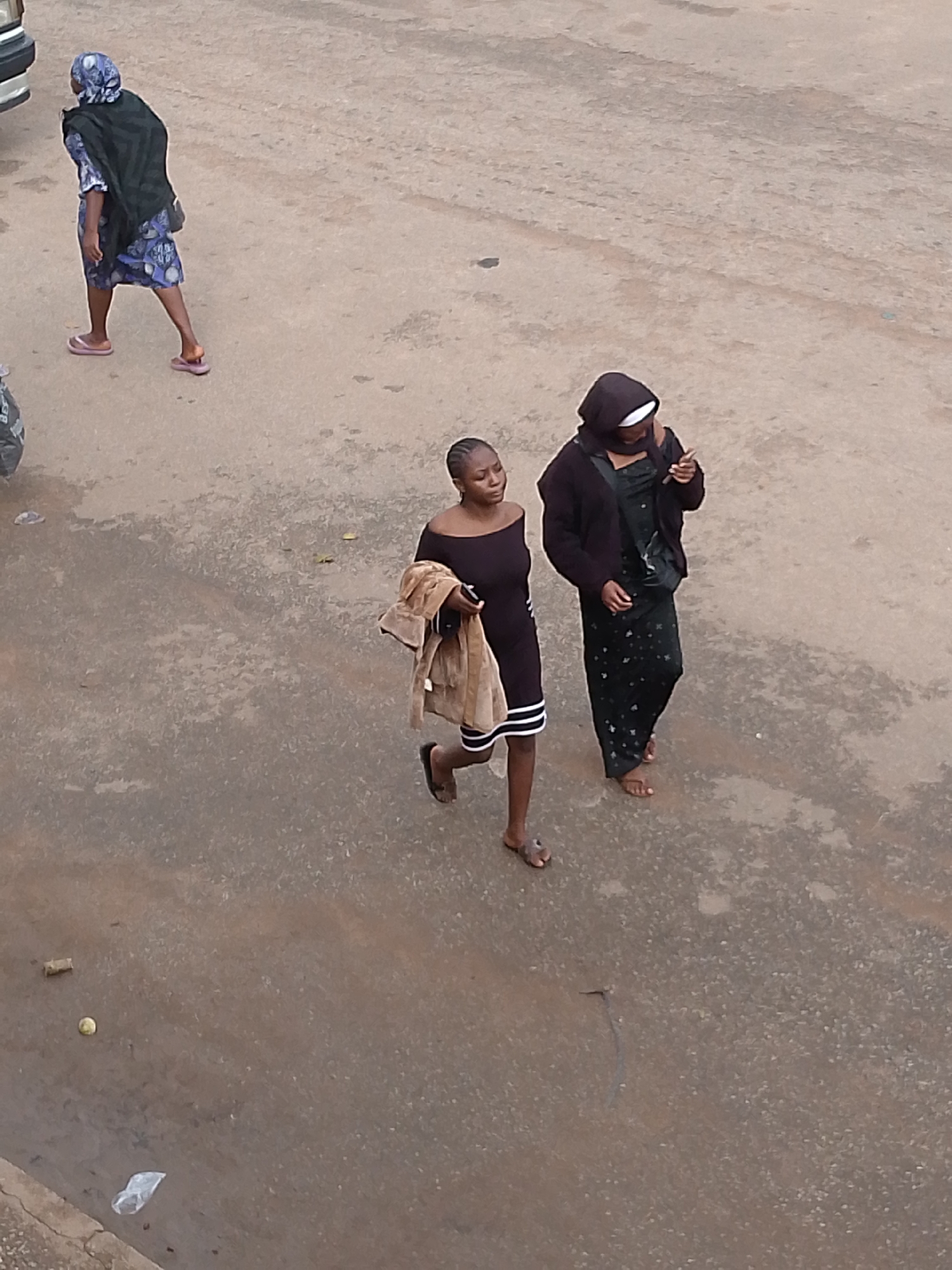 This media file shows three individuals walking on a dirt road. The scene is likely outdoors in a semi-arid or dusty environment, given the ground's appearance. The lighting suggests it could be daytime, possibly overcast or late afternoon, as there are no strong shadows.
The first individual, on the left, is wearing a blue patterned dress and a dark headscarf, with a black garment draped over their shoulders. They are walking away from the camera.
The other two individuals are closer to the center and right of the frame, walking side-by-side. The person on the left is wearing a black off-the-shoulder dress with white stripes on the hem and is carrying a light brown jacket or shawl over their arm. The person on the right is wearing a dark, possibly black, hijab and a dark patterned dress. They are both walking towards the right.
The ground is unpaved and appears to be made of dirt and gravel, with some scattered debris, including a plastic bag and what might be a small fruit or piece of wood. There is a partial view of a vehicle's bumper on the far left of the image.
The overall mood is neutral, with the subjects appearing to be engaged in everyday activity, such as walking to a destination. There is no visible text in the image. The location context of Bukuru, Nigeria, suggests a setting that aligns with the visual cues of a dirt road and the attire of the individuals.