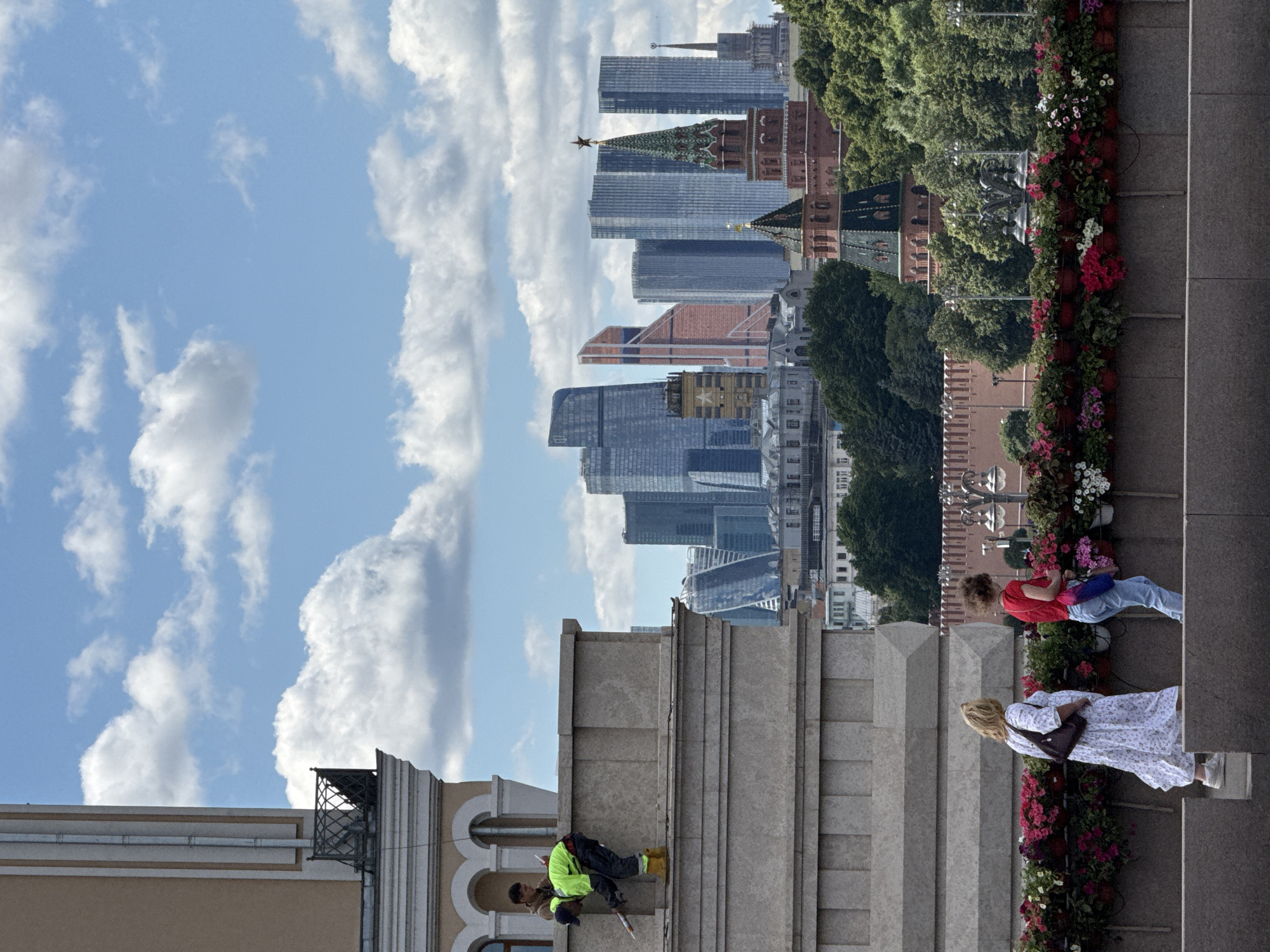 The image captures a view of Moscow, Russia, with the Moscow City skyscrapers in the background. In the foreground, two people are walking along a ledge decorated with flower boxes. One person, wearing a white dress, carries a dark purse. The other person, in a red shirt and jeans, holds a bag and a water bottle.
Further back, on a higher level of a building, two individuals appear to be working. One person is wearing a bright green vest and yellow boots and is leaning over the edge, holding a tool. Another person stands behind them.
The scene is set against a backdrop of blue sky with scattered clouds. In the middle ground, traditional Russian architecture, including a red brick tower with a green spire and a star, is visible amongst lush green trees.