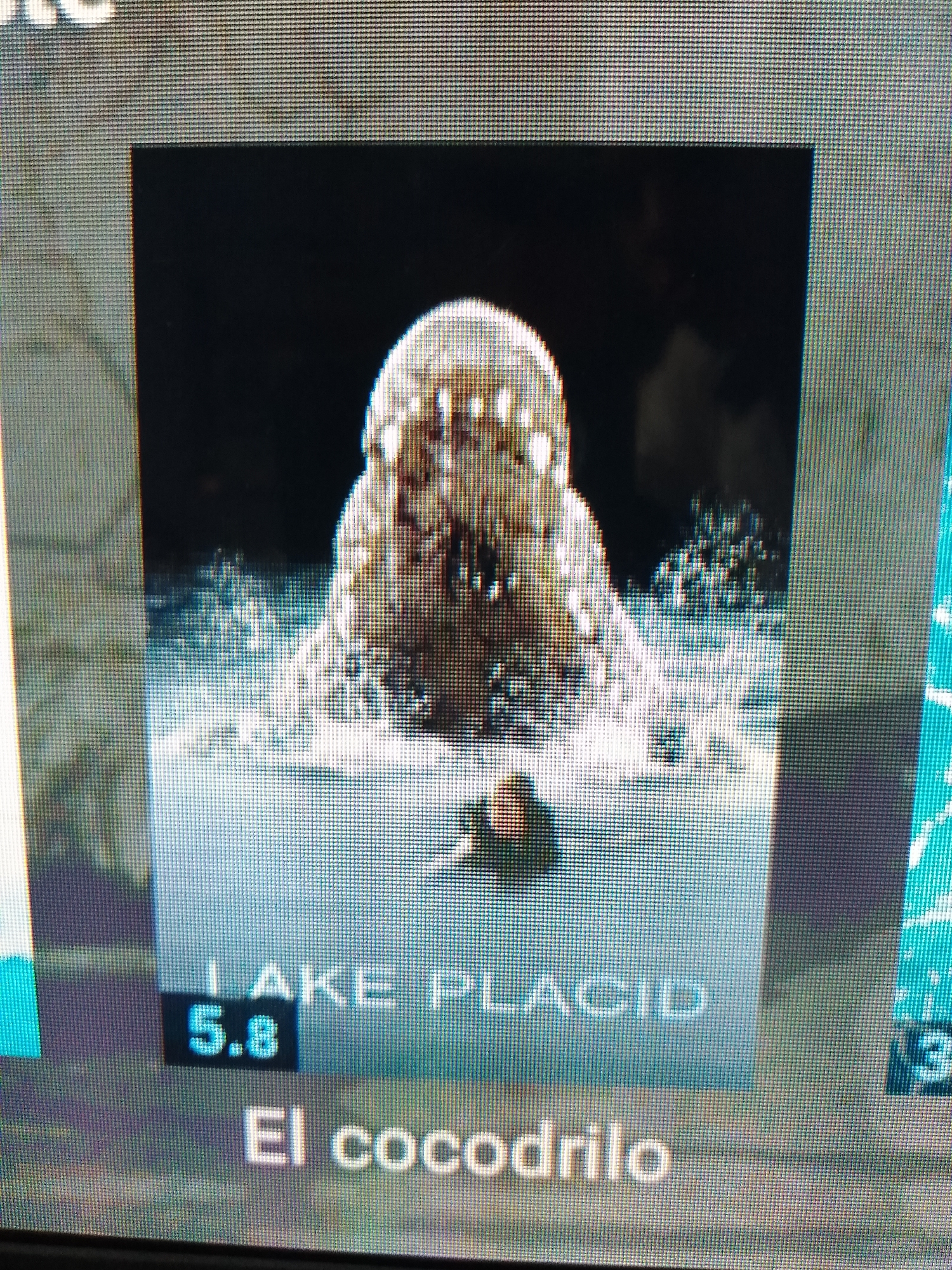 The image shows a television screen displaying the movie poster for "Lake Placid". The poster features a close-up of a crocodile's open mouth emerging from water, with a small boat and a person in the foreground. Text below the image reads "LAKE PLACID" and "5.8" in a black box, followed by "El cocodrilo" in Spanish. The context of Altagracia, Venezuela is not evident in the provided image.