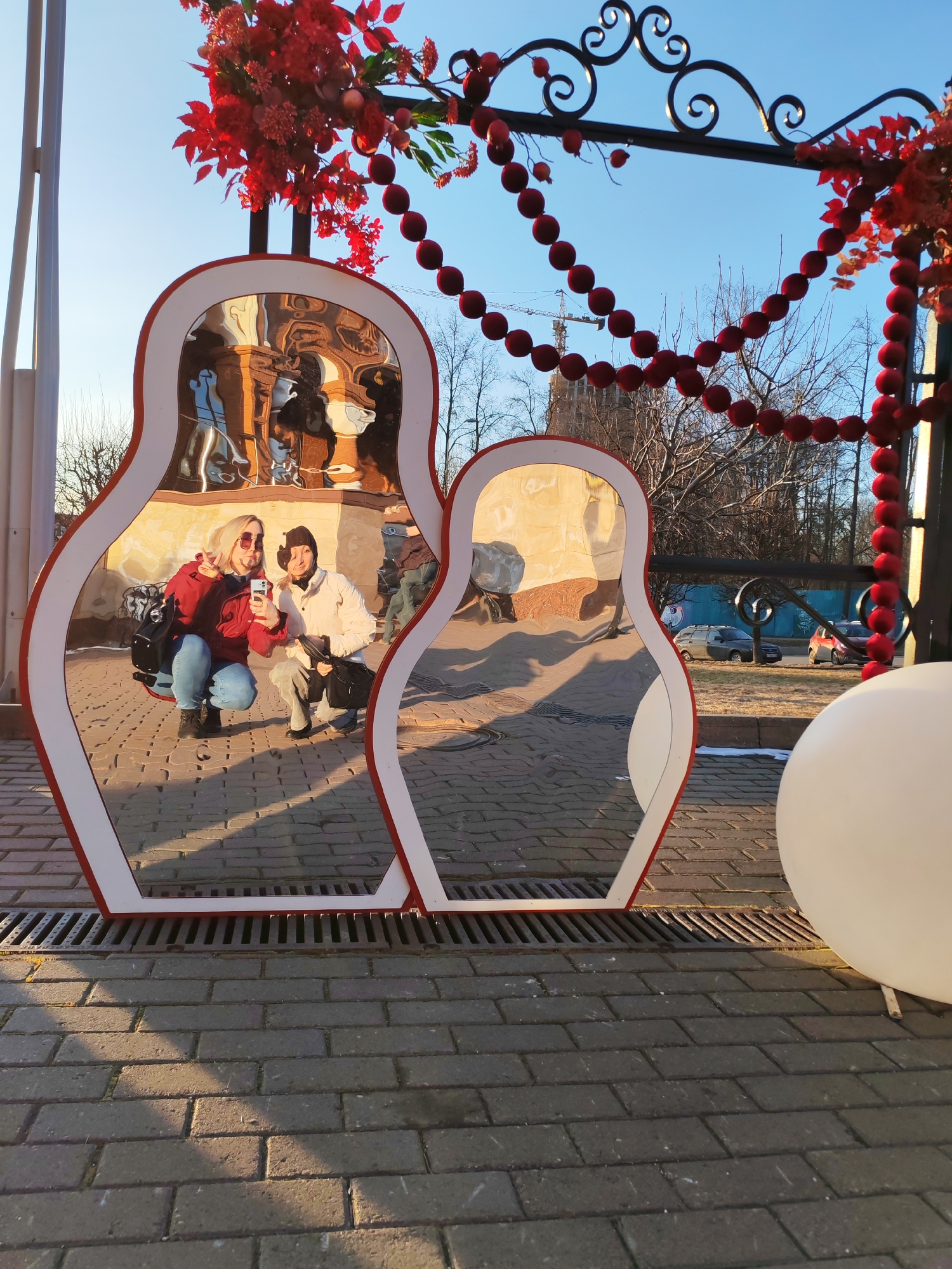 The photograph depicts two women reflected in two matryoshka doll-shaped mirrors. The women are crouching, and one is holding a cell phone as if taking a selfie. The mirrors are framed with red and white trim and are set on a brick-paved surface with a drainage grate underneath. Above the mirrors, there is a decorative archway adorned with red flowers and beads. In the background, outside of the mirrors' reflection, there are buildings, trees, and vehicles. The image was taken in Izhevsk, Russia.