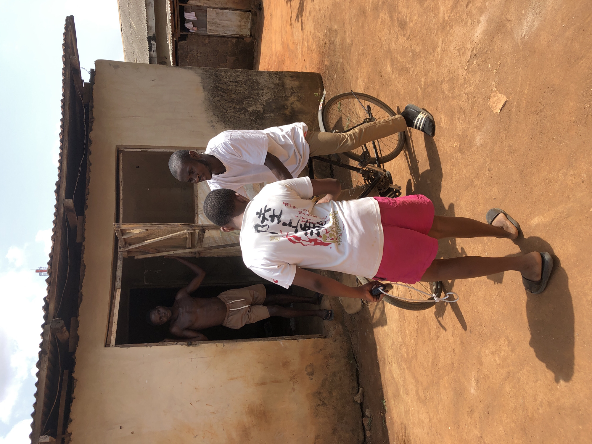 The image shows three individuals near a building in an outdoor setting. One person is inside the window frame of the building, appearing shirtless and wearing shorts. Two other people are standing outside. One is in a white shirt and khaki pants, and the other is wearing a white shirt and pink shorts. The person in khaki pants is looking down, perhaps at something related to the bicycle he is straddling. The person in pink shorts is facing away from the camera, partially obscuring the view of the person inside the window. The setting appears to be an unpaved area with a building that shows signs of age. A bicycle is near the two individuals standing outside. The specific city and location are Unknown.