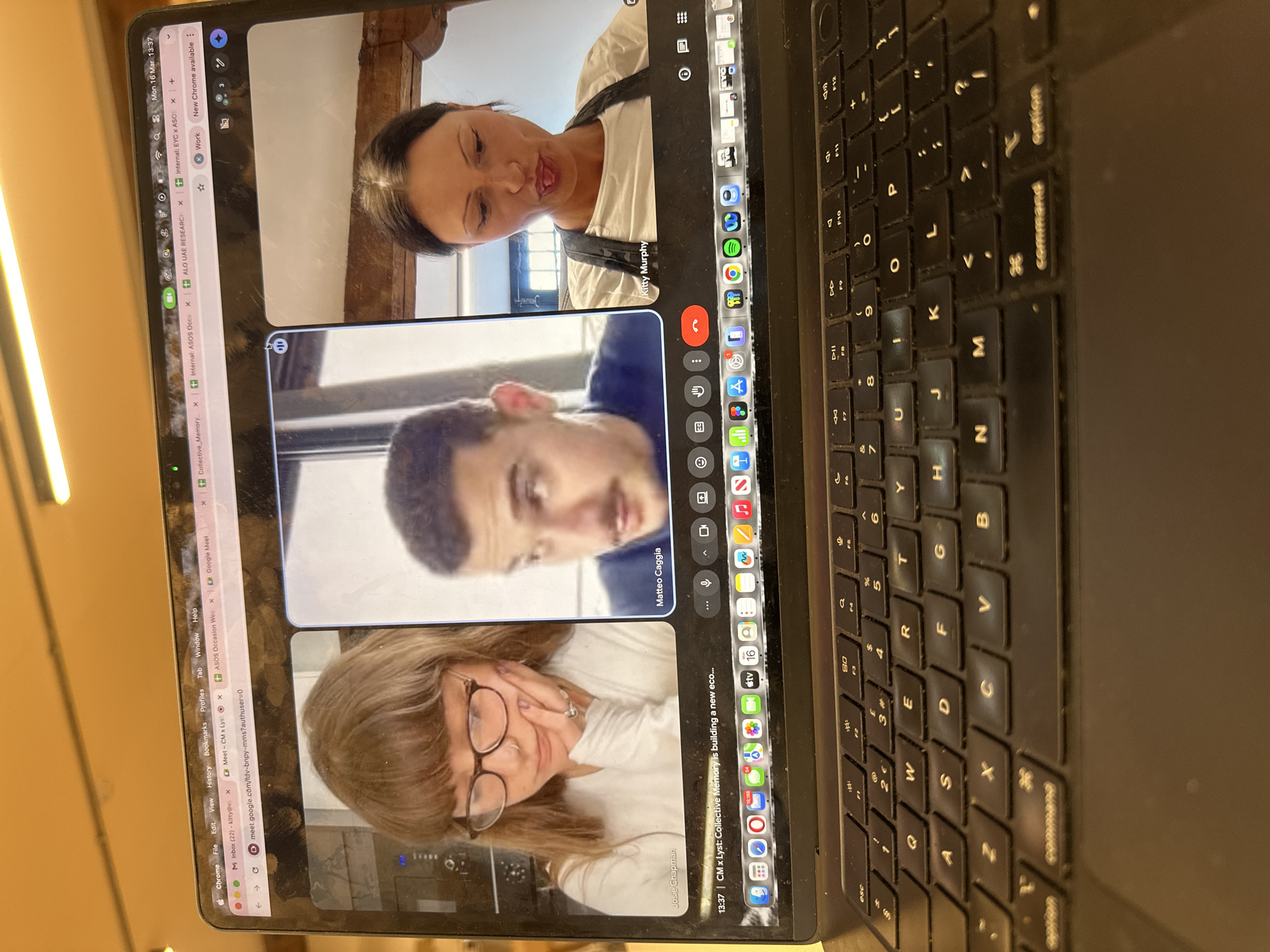 A black MacBook laptop is depicted from a high-angle perspective, displaying an active Google Meet video conference on its screen. The video conference features three participants in distinct panels: a woman identified as Josie Chapman (left, with light brown hair and glasses, resting her hand on her chin), a man identified as Matteo Caggia (center, with short dark hair and a mustache), and a woman identified as Kitty Murphy (right, with dark hair and a dark apron, sticking out her tongue). The meeting title, "CM x Lyst: Collective Memory is building a new eco...", is visible at the bottom of the screen, along with the date and time: "Mon 16 Mar 13:37". Several browser tabs are open, including "Meet - CM x Lys" and tabs referencing "ASOS Occasion Wea" and "Internal: EYC x ASOS". Given that ASOS and Lyst are prominent British companies headquartered in London, the scene is contextually located in London, United Kingdom. The laptop's QWERTY keyboard is visible in the foreground, displaying dust accumulation and some wear on the keys. The background above the laptop is blurred, showing warm, artificial overhead lighting in an indoor setting.