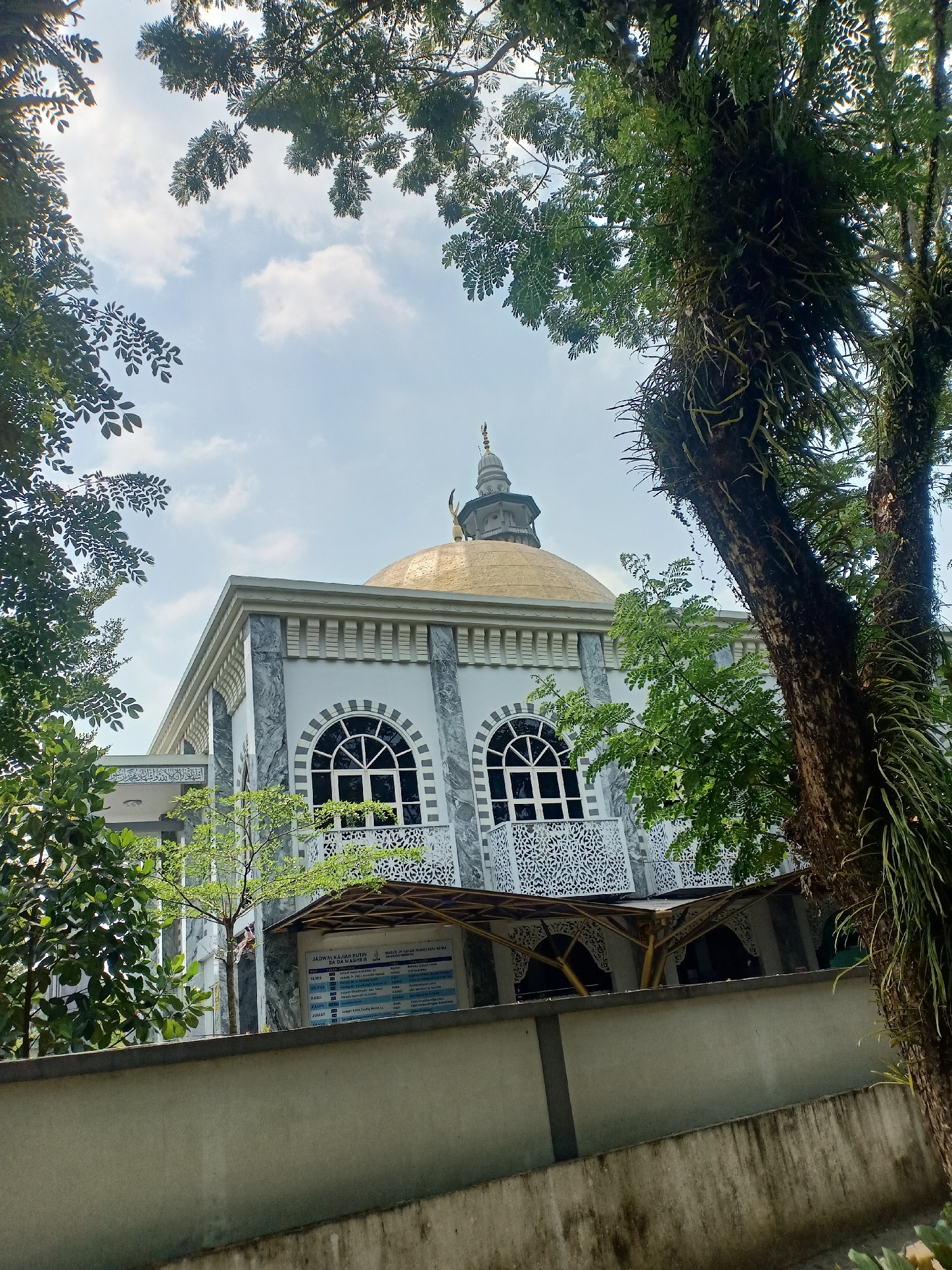 This image captures a mosque with a prominent golden dome, surrounded by lush green trees and a bright blue sky with scattered clouds. The mosque features elegant architecture with white marble accents and arched windows. A grey wall runs in front of the building, with some greenery peeking over it. To the right, a large tree with moss and hanging roots adds a natural, textured element to the scene. A sign is visible on the wall, displaying text in Indonesian, likely providing information about the mosque's schedule. The overall atmosphere is serene and tranquil, suggesting a peaceful urban setting in Semarang, Indonesia.