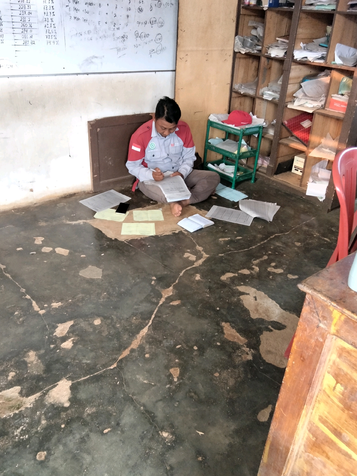In a dimly lit room, a man wearing a light-colored uniform with red accents sits on the floor, engrossed in paperwork. He is surrounded by scattered documents and a smartphone. To his right, a wooden shelf unit is filled with files and various items. A red chair is partially visible in the background. The floor is concrete with visible cracks and patches of discolored material. The scene appears to be inside a building in Teluk Tenggirik, Indonesia.